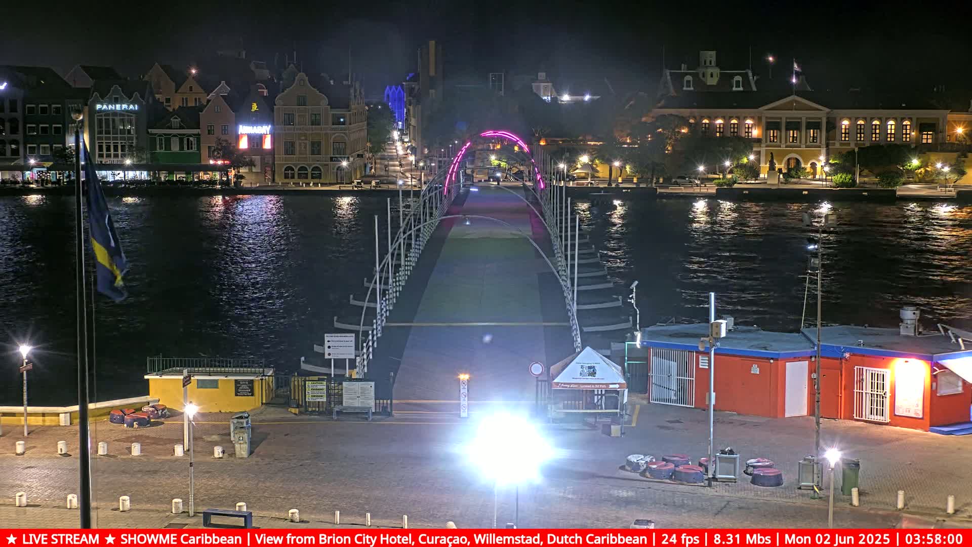 A nighttime view of a brightly lit pedestrian bridge spanning a calm waterway, with colorful buildings lining the shore.