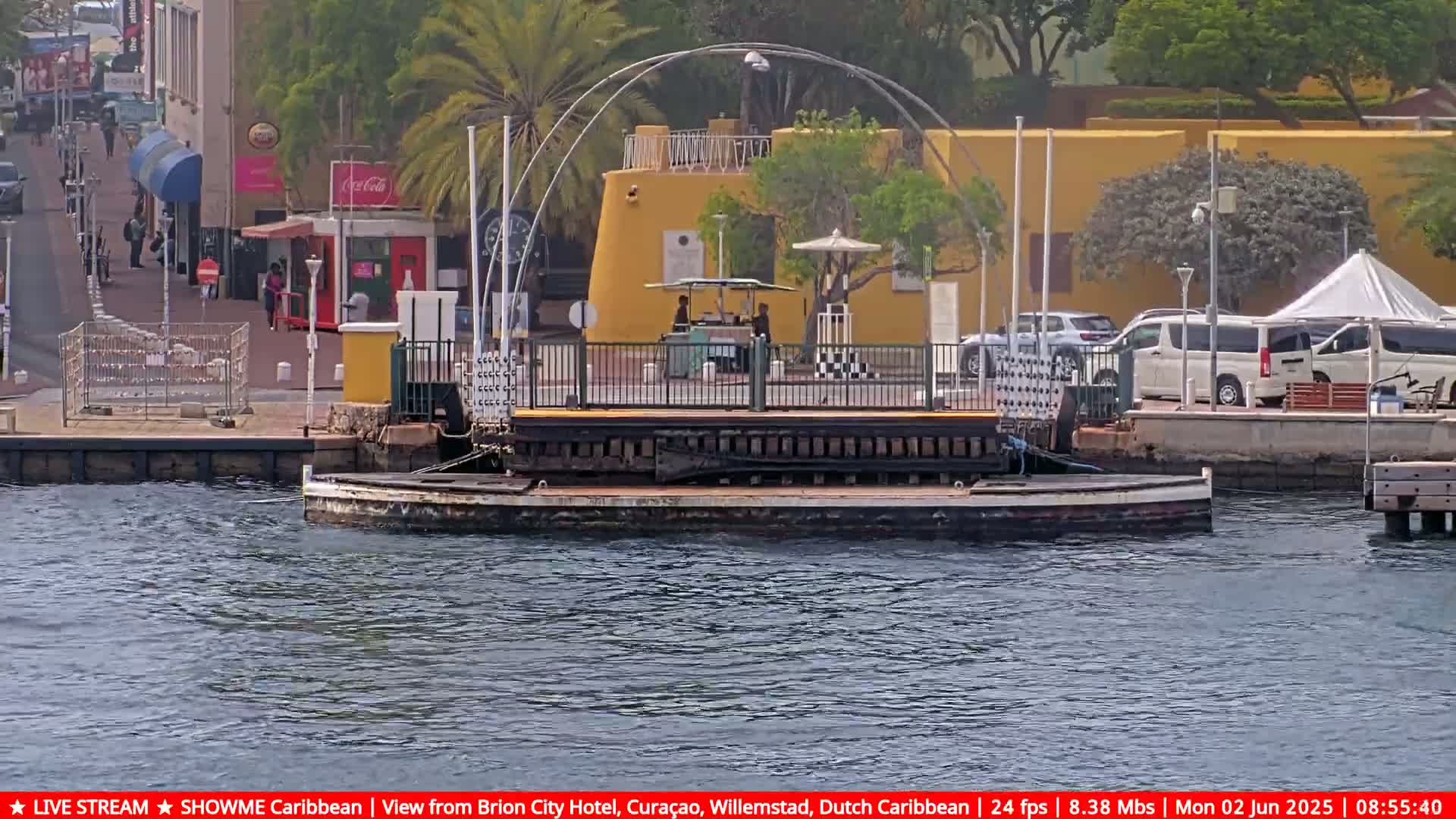 A floating pontoon bridge sits in calm water near a yellow building and a waterfront area with some people and parked vehicles.