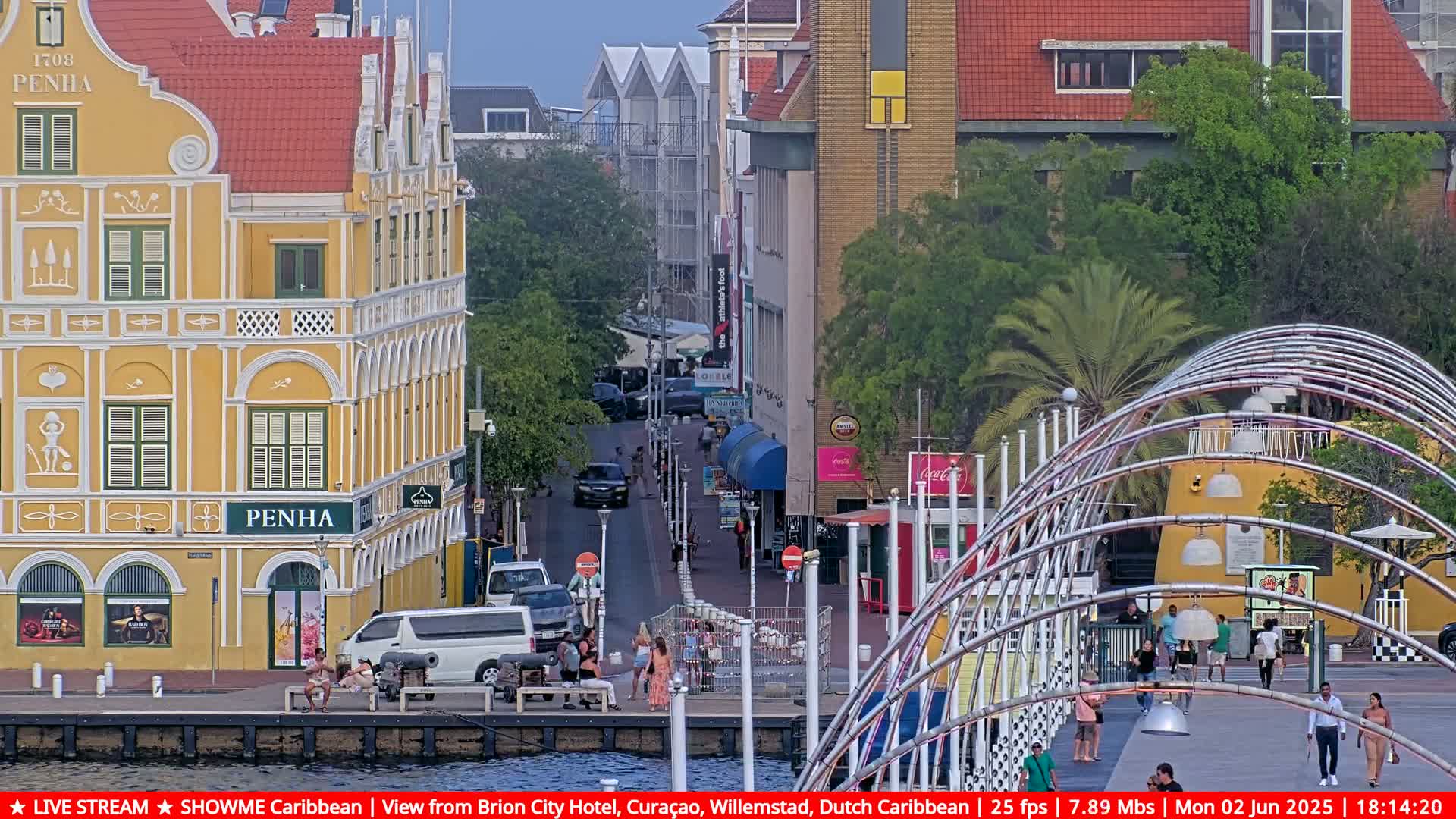 A street scene in a waterfront city with colorful colonial-style buildings, under an overcast sky, shows people walking and sitting near a curved metal walkway.