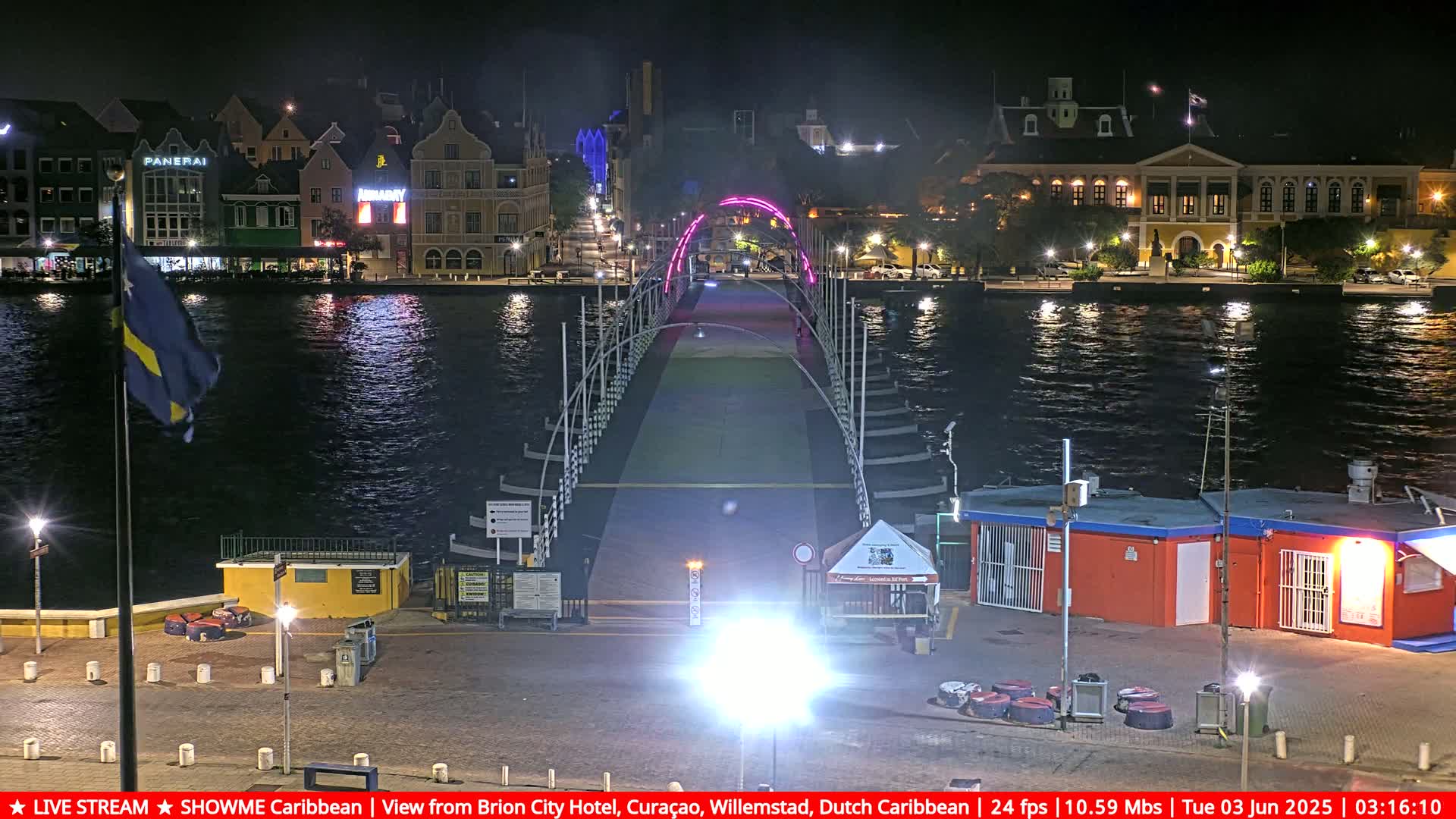 A nighttime view of a pink-lit floating bridge spanning a waterway, with colorful buildings and a flagpole on either side.