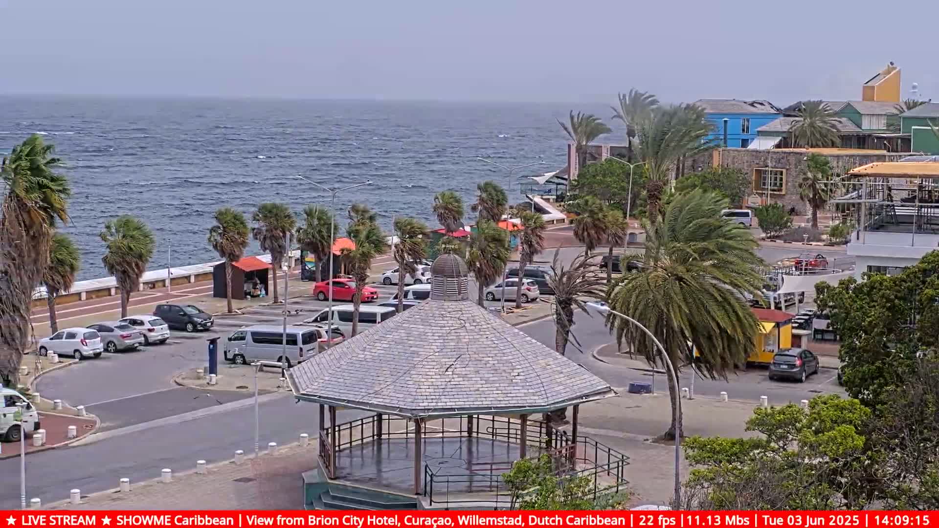 A seaside parking lot with palm trees and a gazebo is shown on an overcast day with a choppy ocean in the background.