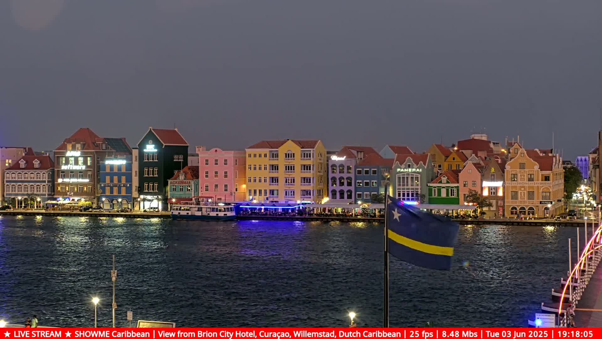 A row of brightly lit, colorful buildings lines a waterfront at dusk, with a Curaçao flag visible in the foreground.