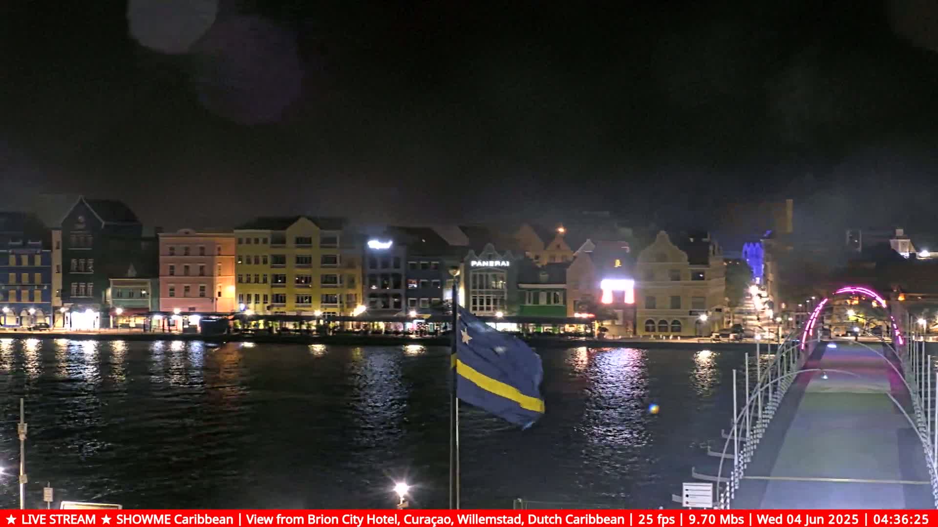 A nighttime view of a waterfront city, illuminated with colorful buildings and lights, featuring a flag and a bridge spanning a calm body of water under a dark sky.