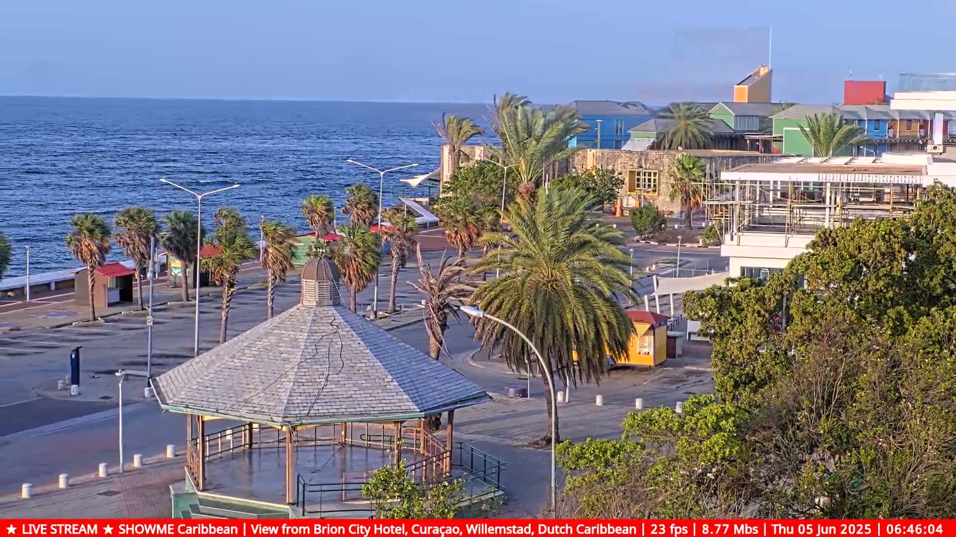 A seaside plaza with palm trees, a gazebo, and colorful buildings under a mostly clear sky, overlooking a calm ocean.