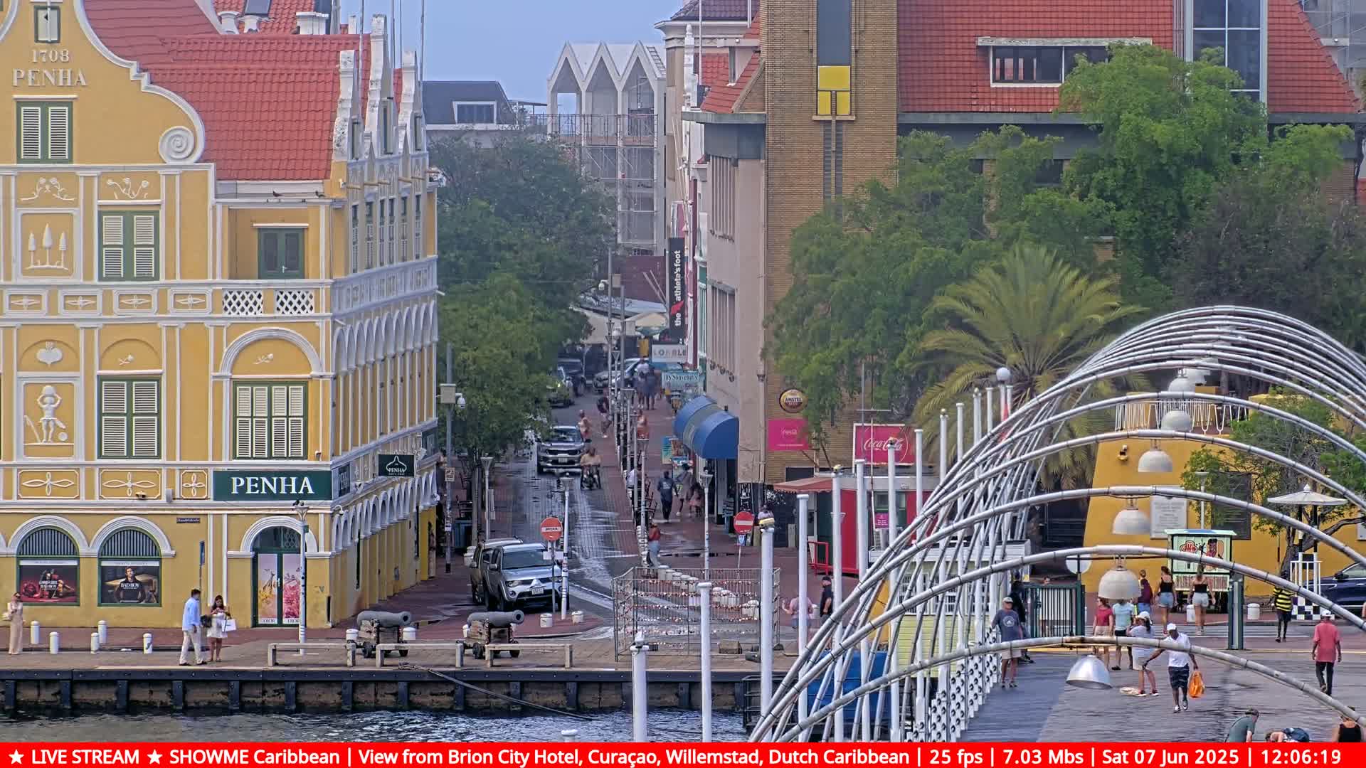 A wet, paved street lined with colorful buildings, some with ornate detailing, leads to a modern, arched pedestrian bridge under an overcast sky.
