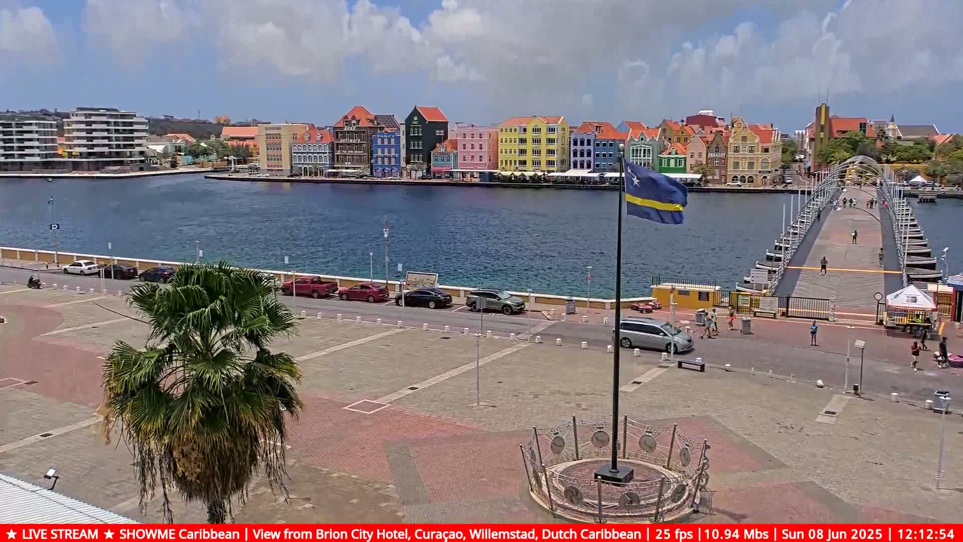 A partly sunny day shows a waterfront scene with colorful buildings, a pontoon bridge, parked cars, and a palm tree, all viewed from an elevated position.