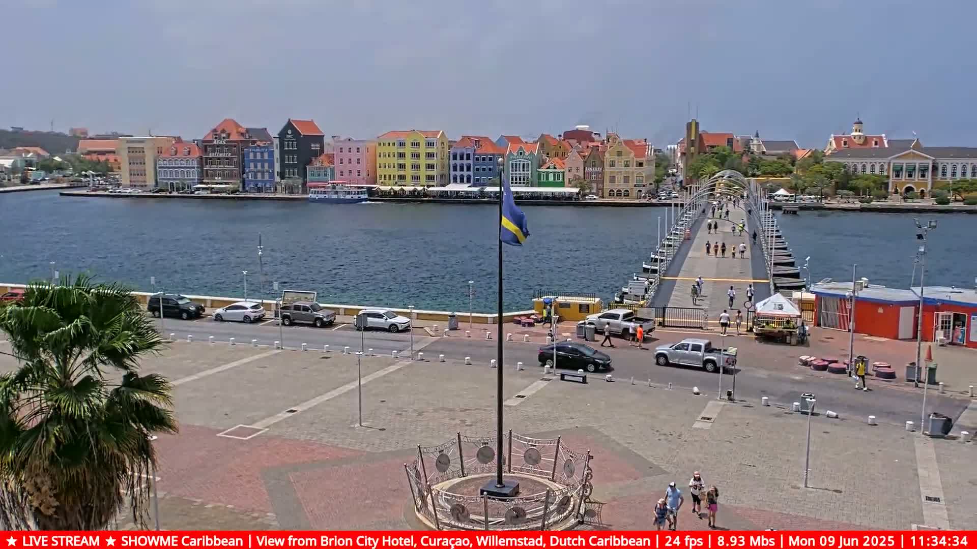 A sunny day shows a waterfront scene with colorful buildings, a floating bridge with pedestrians, and cars parked in a plaza, all under a clear sky.