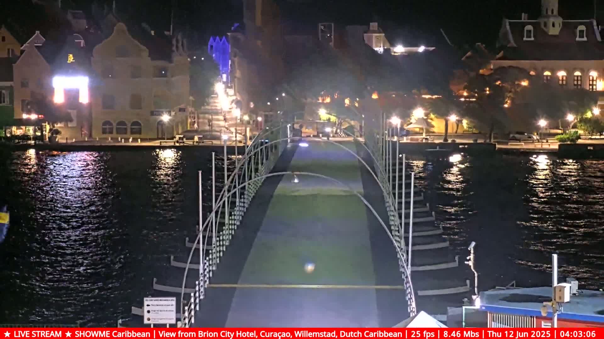 A nighttime view of a bridge over calm water, with brightly lit buildings lining the waterfront on either side.