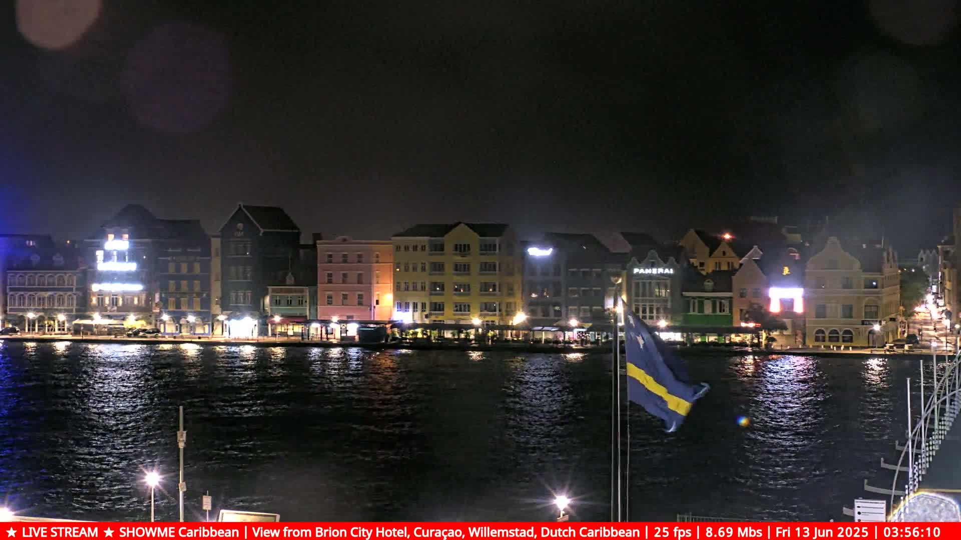 A nighttime waterfront scene shows a row of brightly lit buildings along a dark body of water, with a flag waving gently in the breeze.