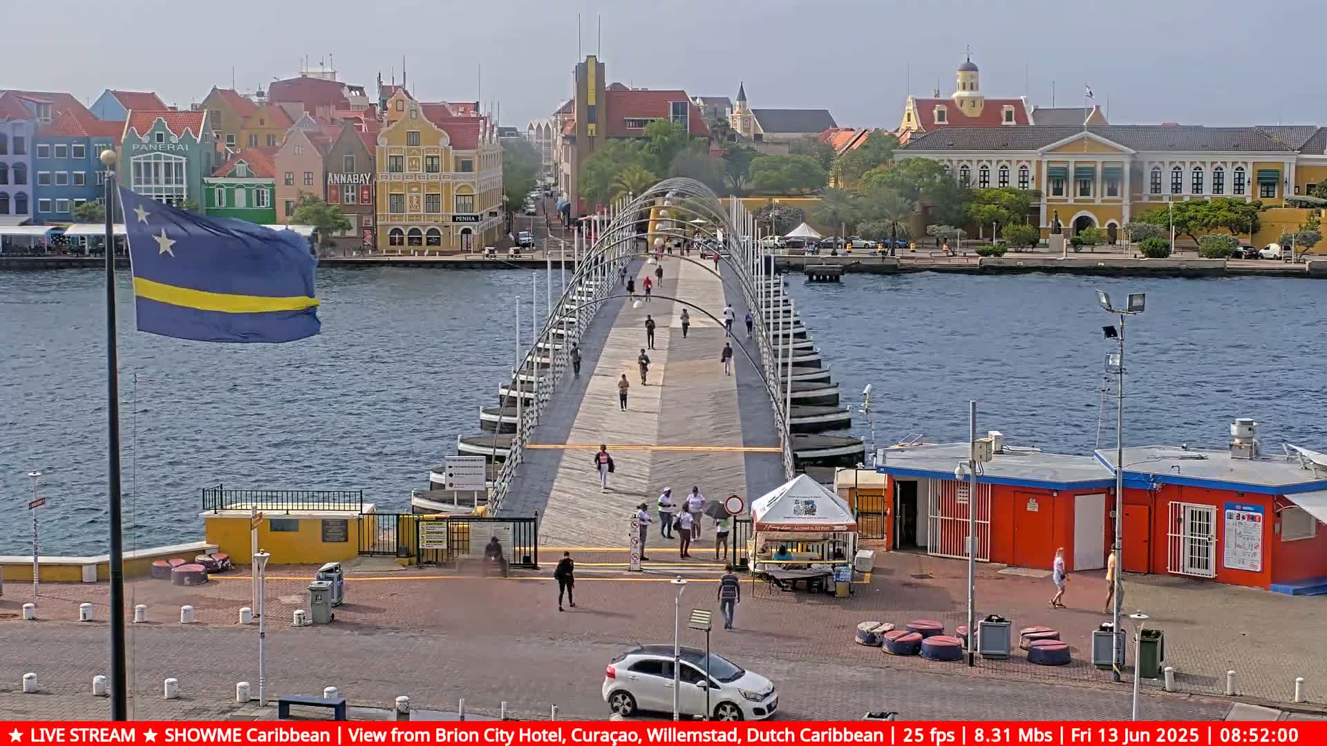 A pontoon bridge with pedestrians spans a waterway, with colorful buildings and a Curaçao flag in the background under a sunny sky.
