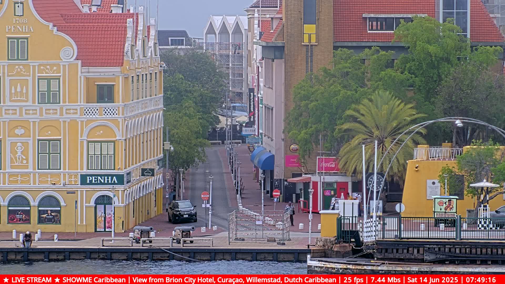 A partly cloudy day reveals a waterfront street scene with colorful colonial buildings, a few cars, and a temporary barrier closing off the road.