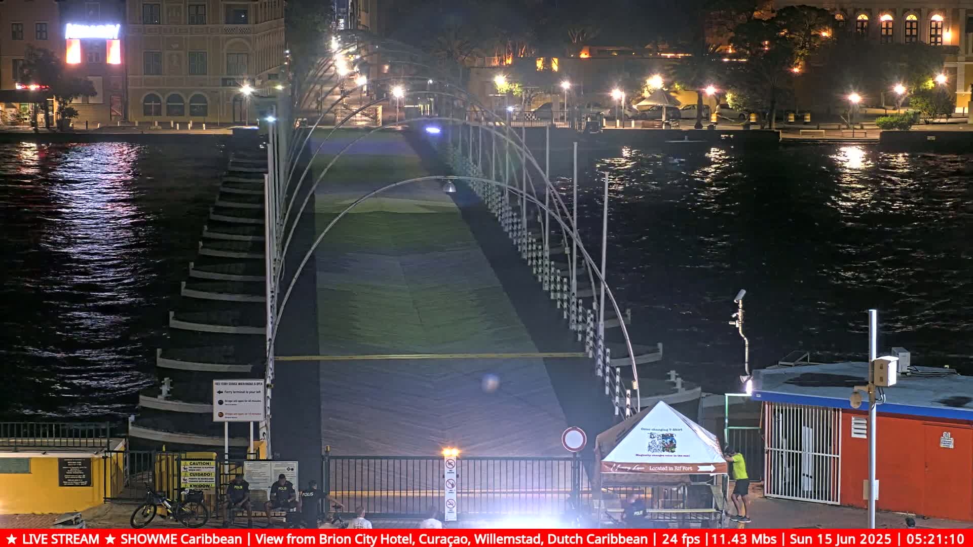 A long, arched pedestrian bridge stretches across a dark, gently moving body of water at night, illuminated by lights along the bridge and buildings on the shore.