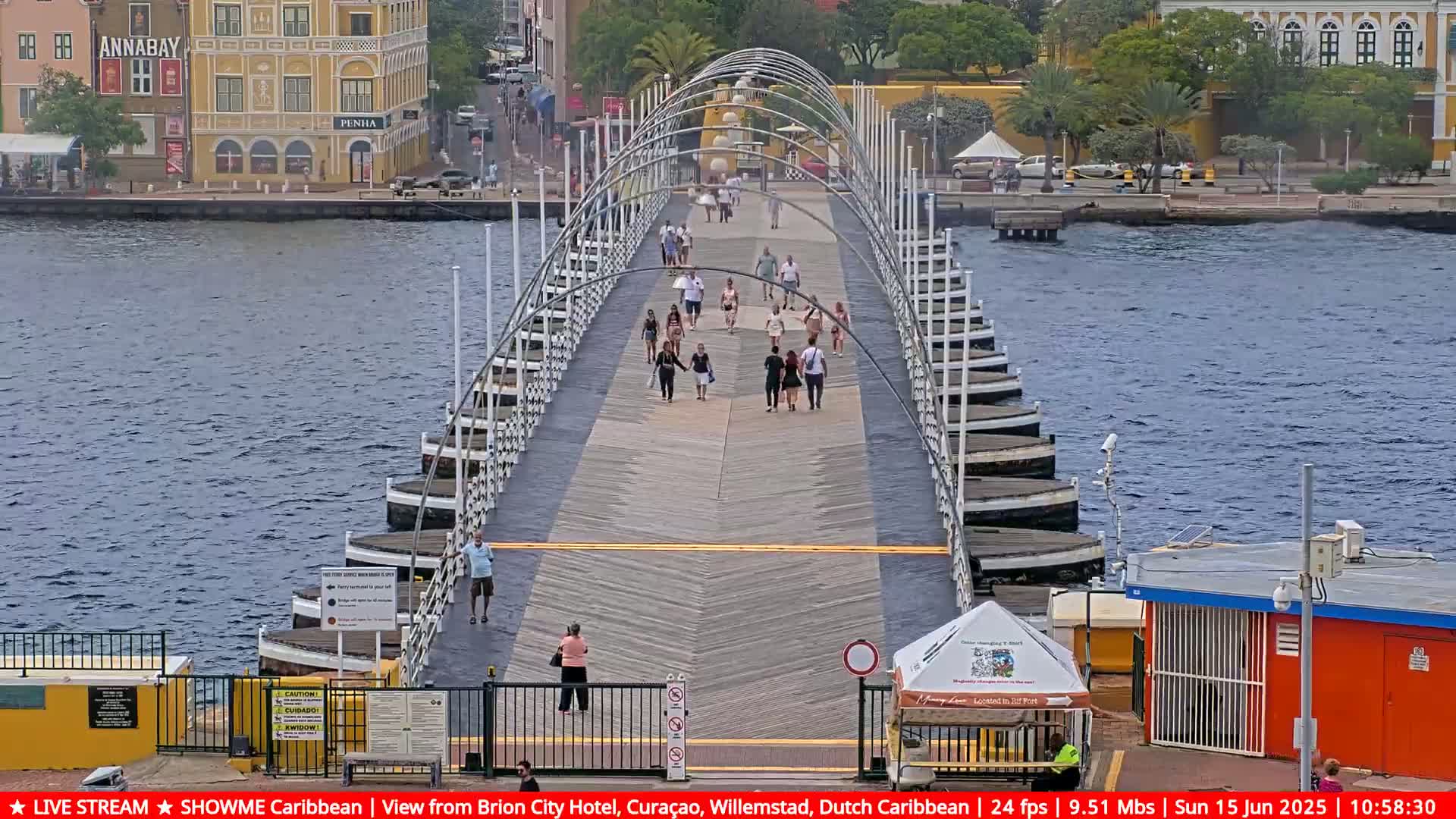 A pontoon bridge with pedestrians stretches across a body of water, under a partly cloudy sky, with colorful buildings visible on the far shore.