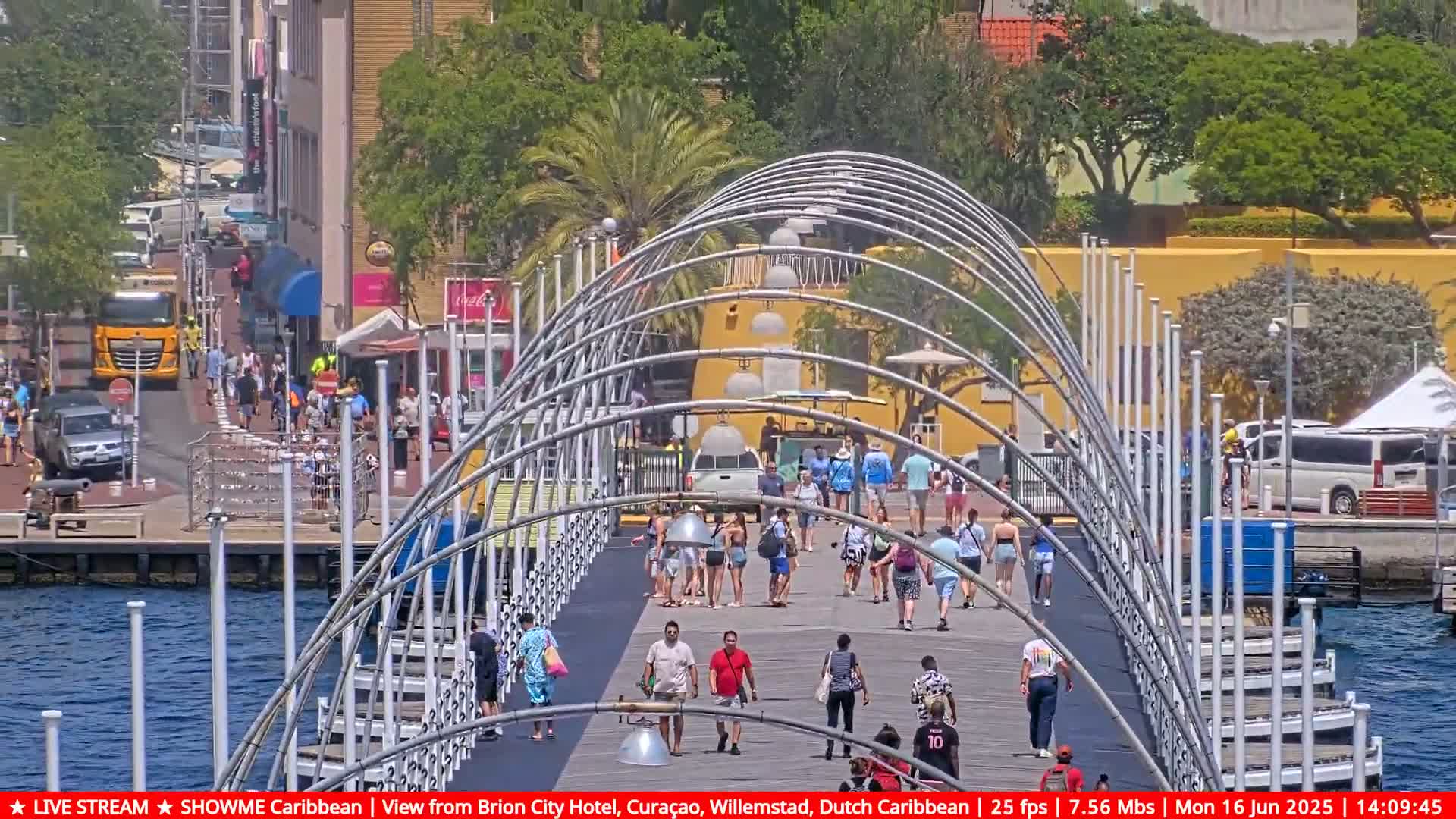 A pedestrian bridge with a curved metal framework spans a waterway, filled with people walking under a sunny sky.
