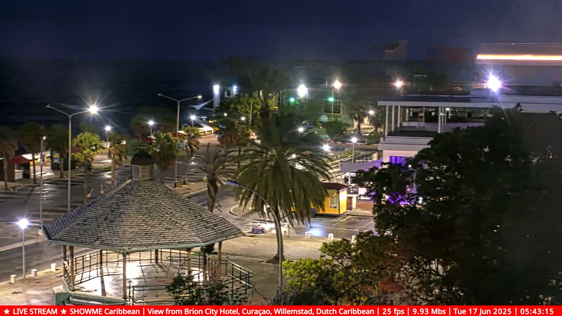 A nighttime view of a coastal town square with a gazebo in the foreground, palm trees, and buildings under a dark sky.