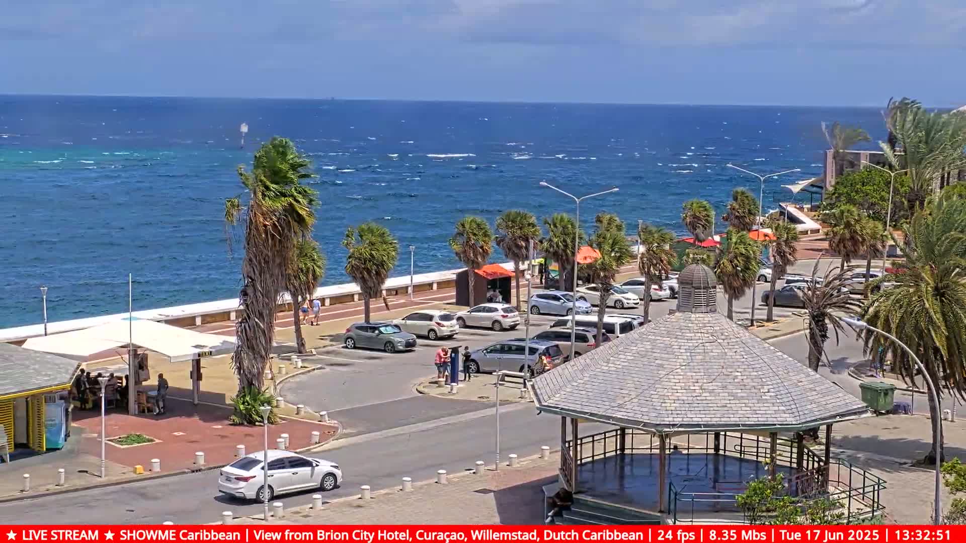 A sunny seaside parking lot with palm trees, a gazebo, and a few cars, overlooks a blue ocean with small waves.