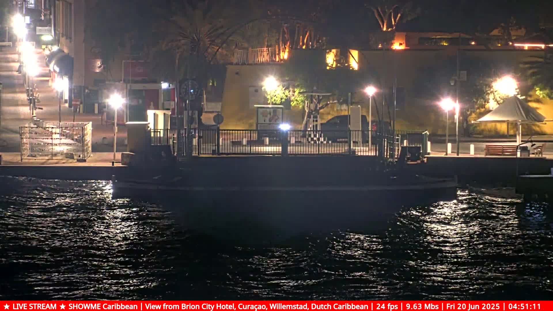 A nighttime view of a waterfront area with a small dock, illuminated buildings, and slightly choppy water.