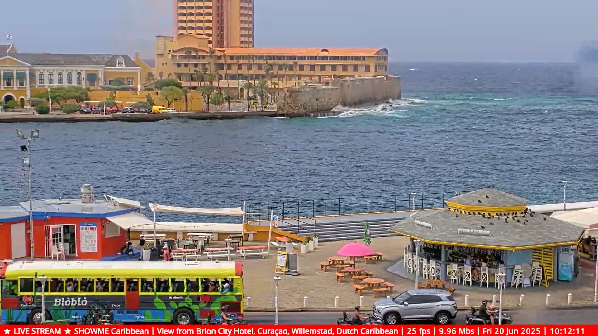 A waterfront scene shows a yellow bus, several buildings, and an outdoor eatery under a partly cloudy sky with choppy water.