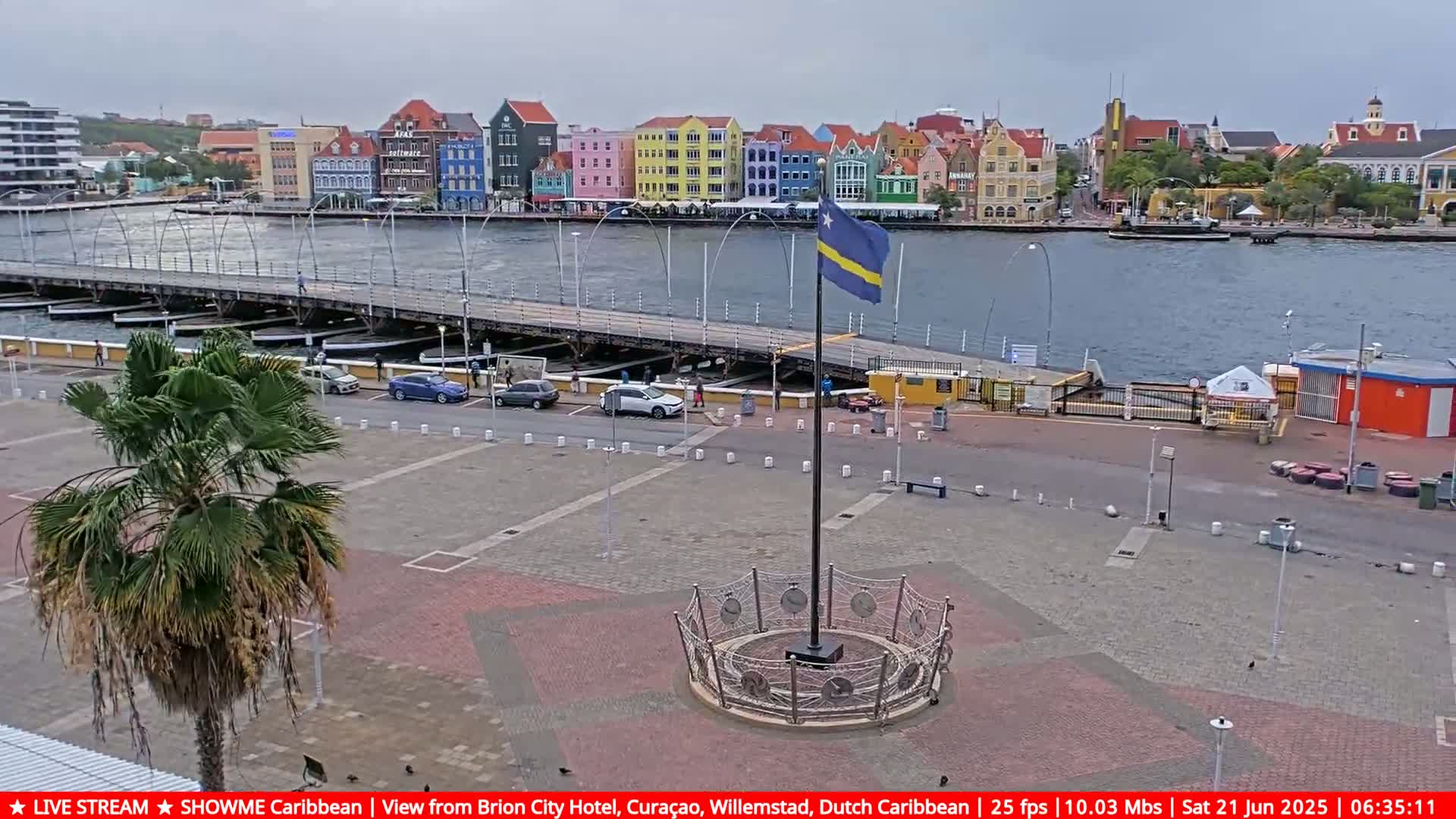 A waterfront plaza with a flagpole and a circular monument, overlooked by colorful buildings and a floating bridge, under an overcast sky.