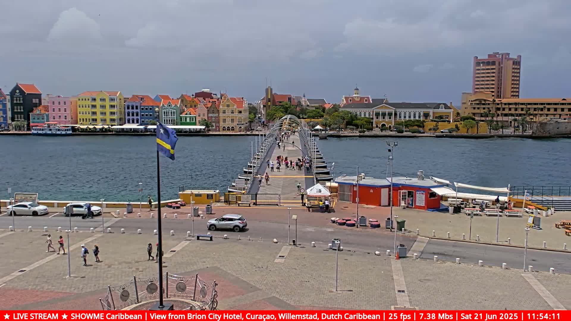 A pontoon bridge stretches across a calm body of water, connecting a colorful waterfront city to a plaza with several pedestrians and parked cars under a partly cloudy sky.