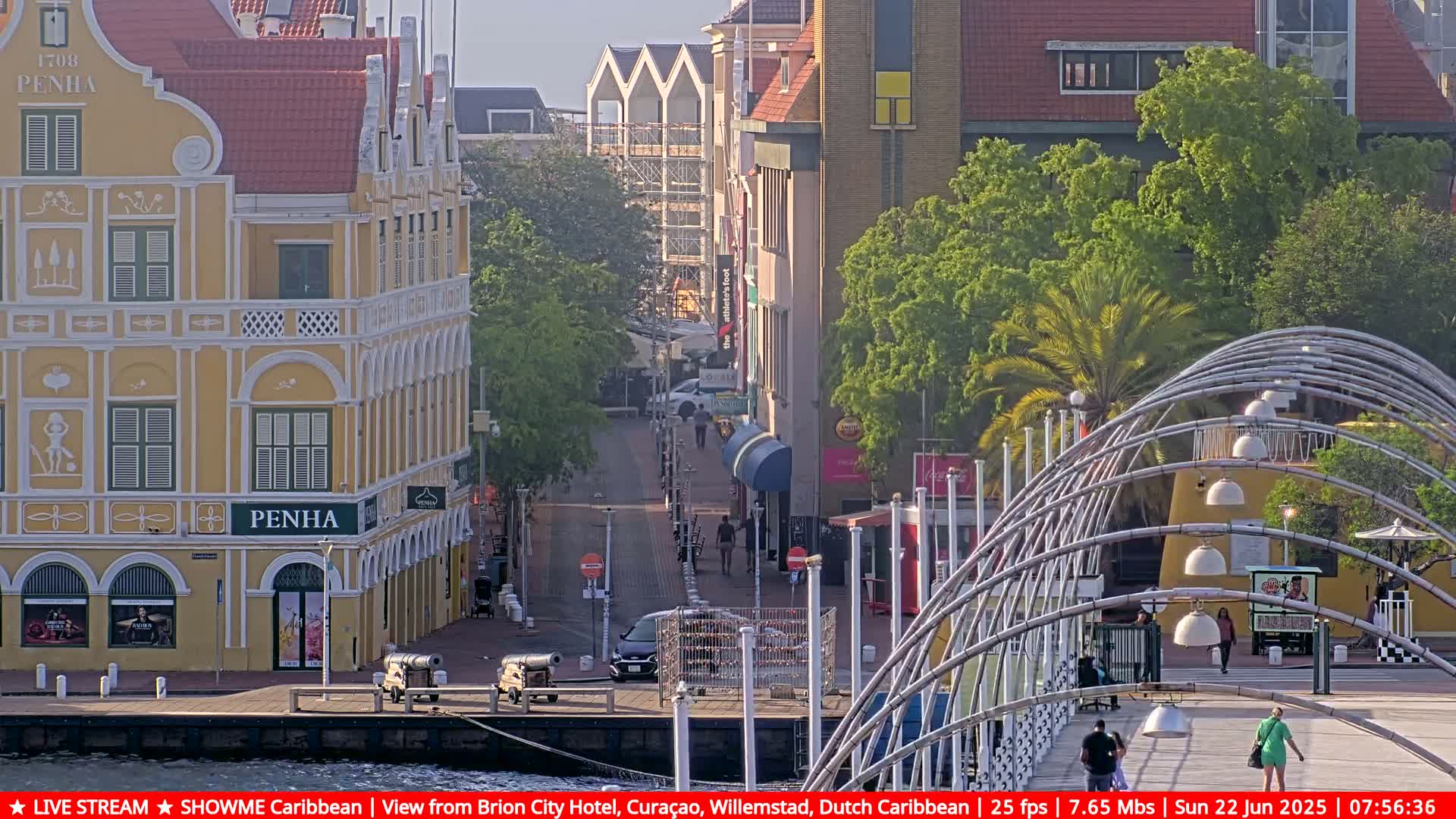 A sunny waterfront street scene shows pedestrians walking past colorful colonial-style buildings, with a modern arched bridge in the foreground.