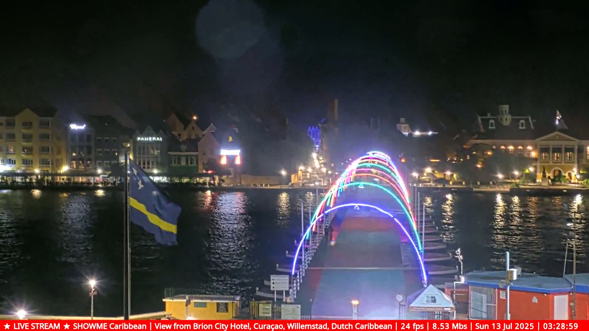 A brightly lit bridge with multicolored arches spans a waterway at night, with city buildings illuminated along the shore and a flagpole displaying a flag in the foreground.