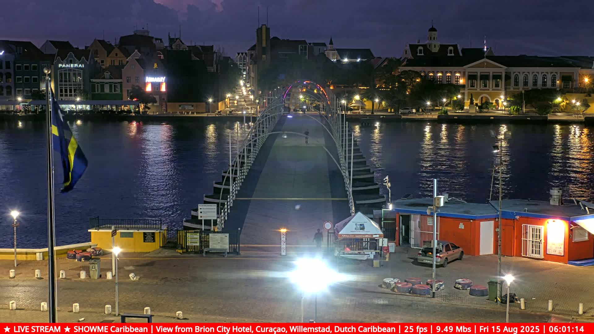 A nighttime waterfront scene with rain-streaked glass obscuring the view of a shoreline with outdoor seating and small buildings, illuminated by artificial lights.