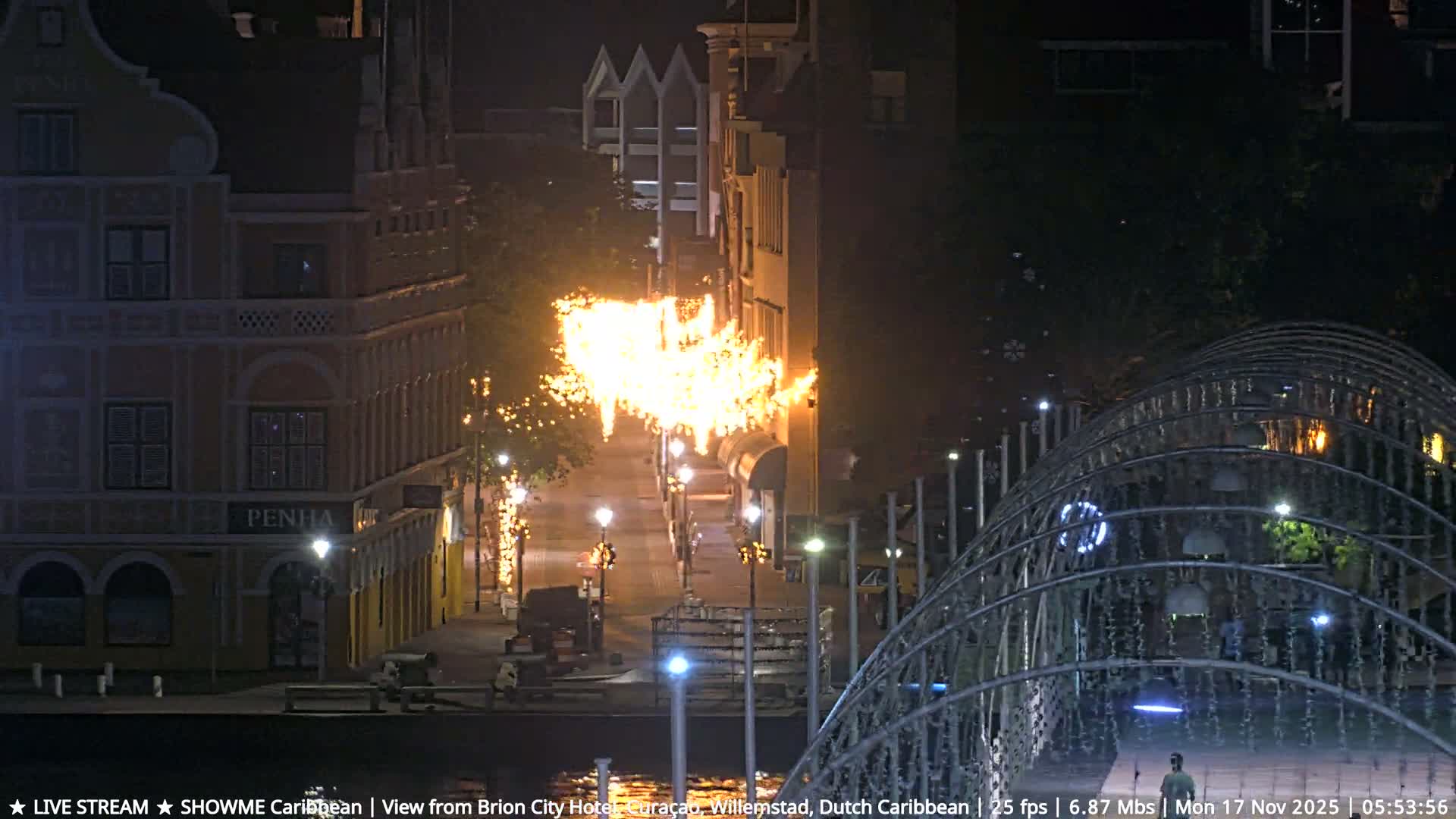 Large, bright flames erupt along a street lined with buildings and decorative lights at night, with a waterfront visible on the left and a metal archway on the right, under clear skies.