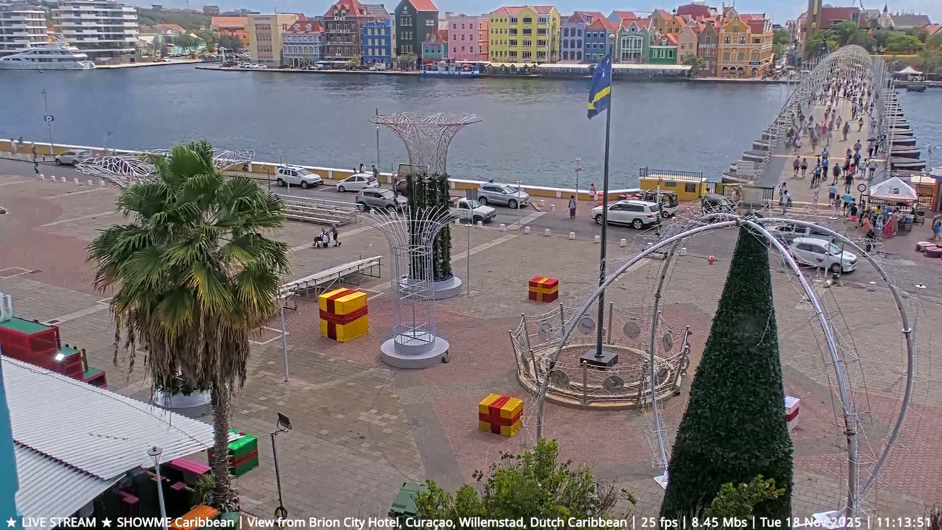 Large, bright flames erupt along a street lined with buildings and decorative lights at night, with a waterfront visible on the left and a metal archway on the right, under clear skies.