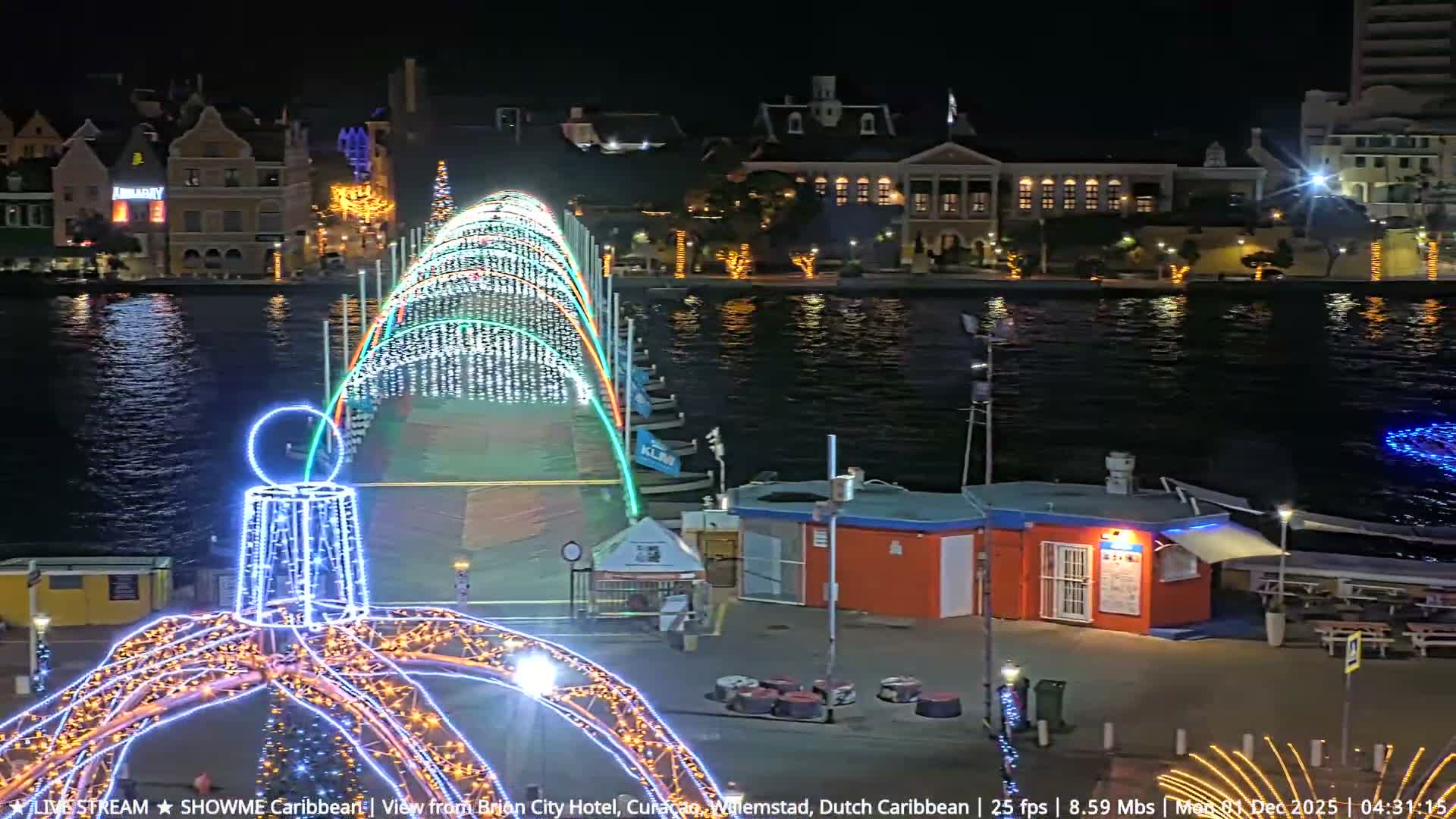 A clear night illuminates a city waterfront, featuring a large bridge spectacularly adorned with white and multicolored festive lights spanning dark water, with similarly lit buildings reflecting across the calm surface and more decorative lights on a foreground dock.