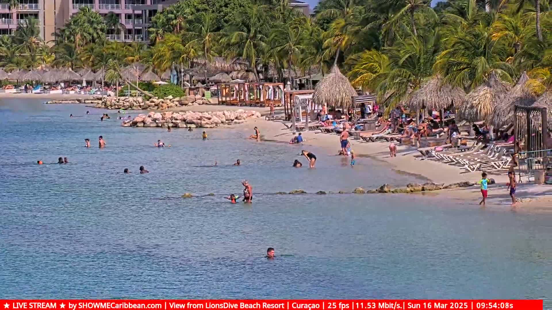 A sunny beach scene shows many people swimming and relaxing on the sand under thatched-roof beach umbrellas near a resort.