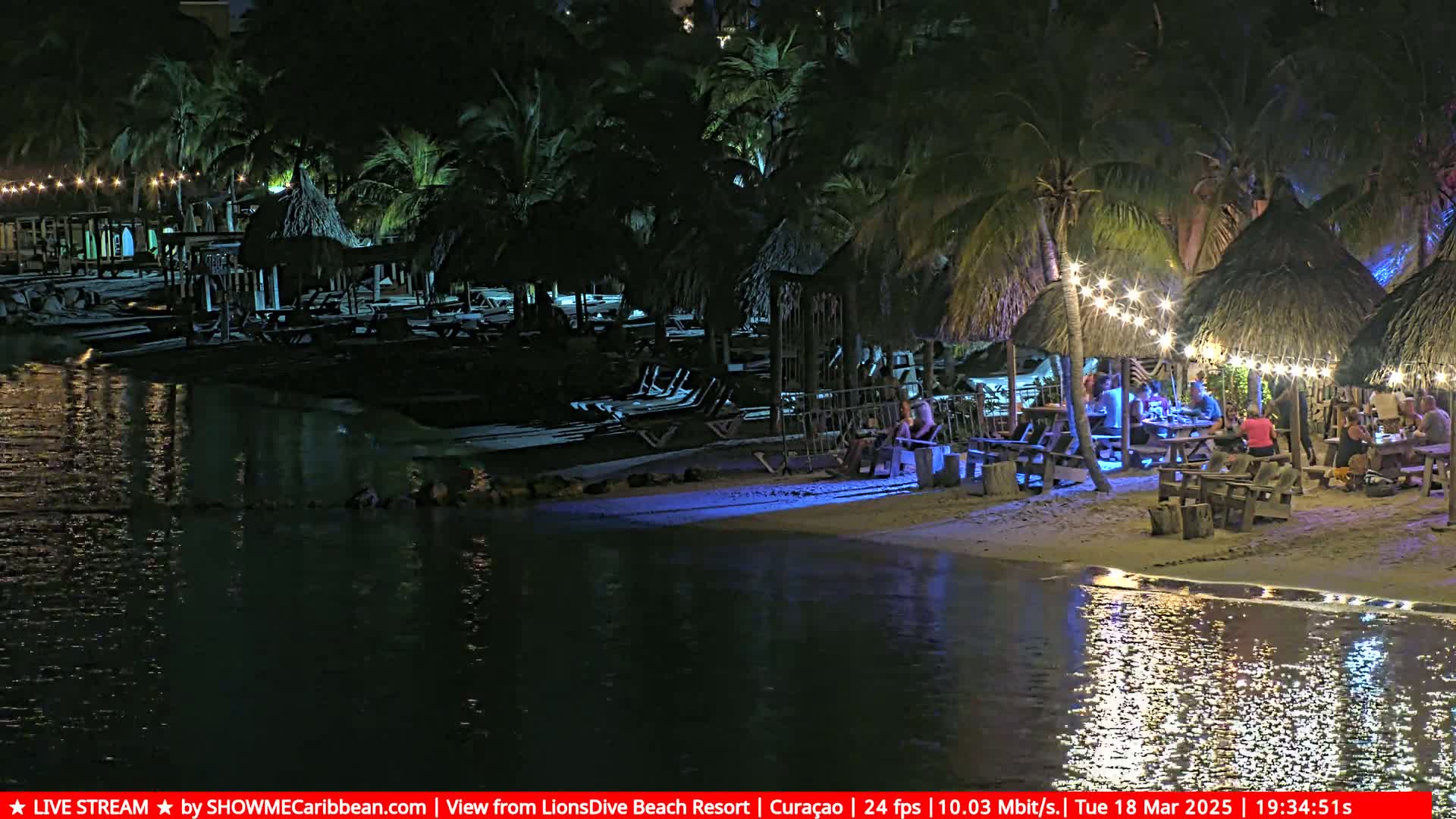 A nighttime beach scene shows a calm body of water reflecting lights from thatched-roof structures where people are seated at tables under string lights.