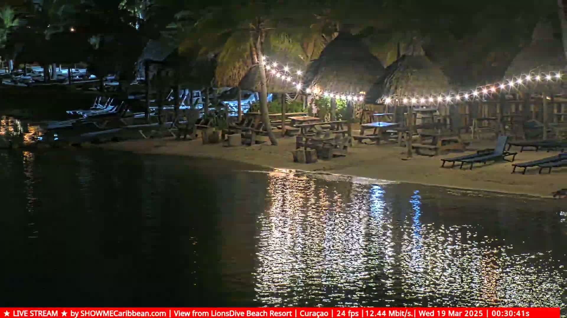 A nighttime beach scene shows several thatch-roofed structures with string lights, picnic tables, and lounge chairs situated on a sandy shore next to calm water.