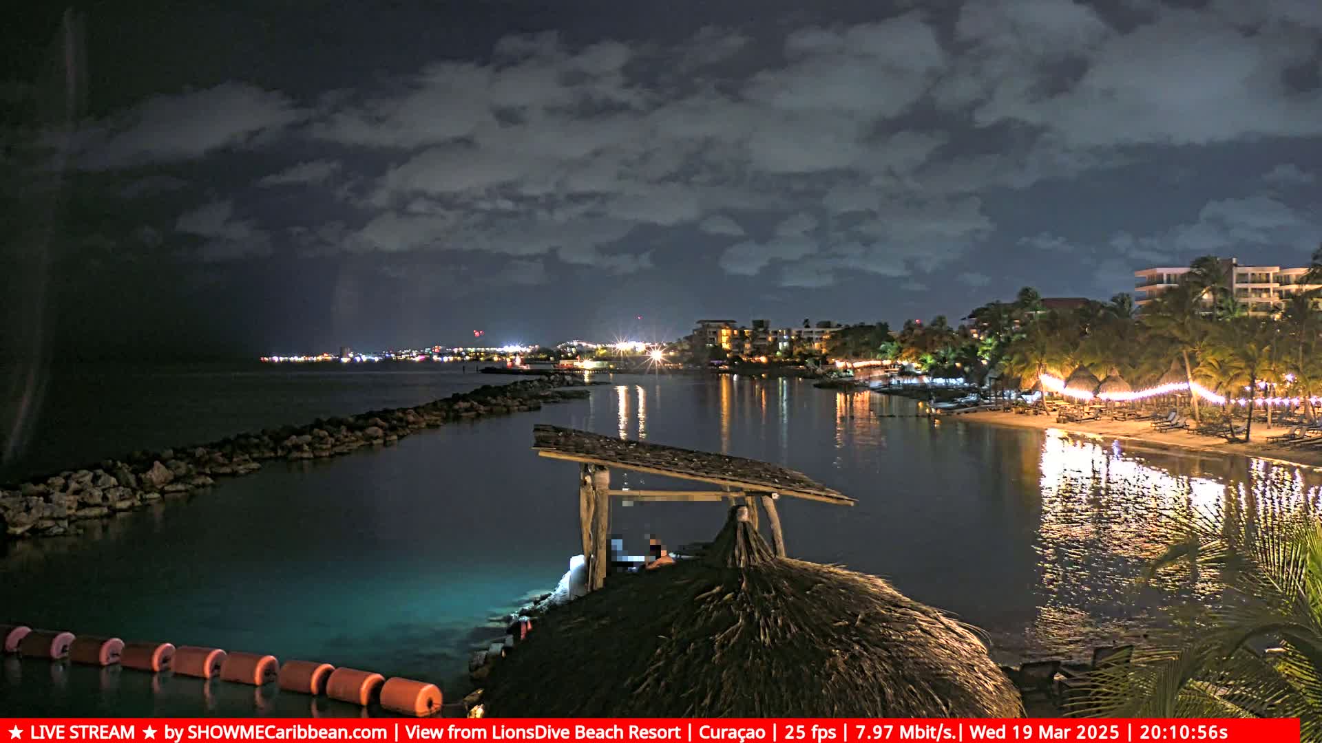 A nighttime view of a calm, illuminated bay with a resort shoreline under a cloudy sky.