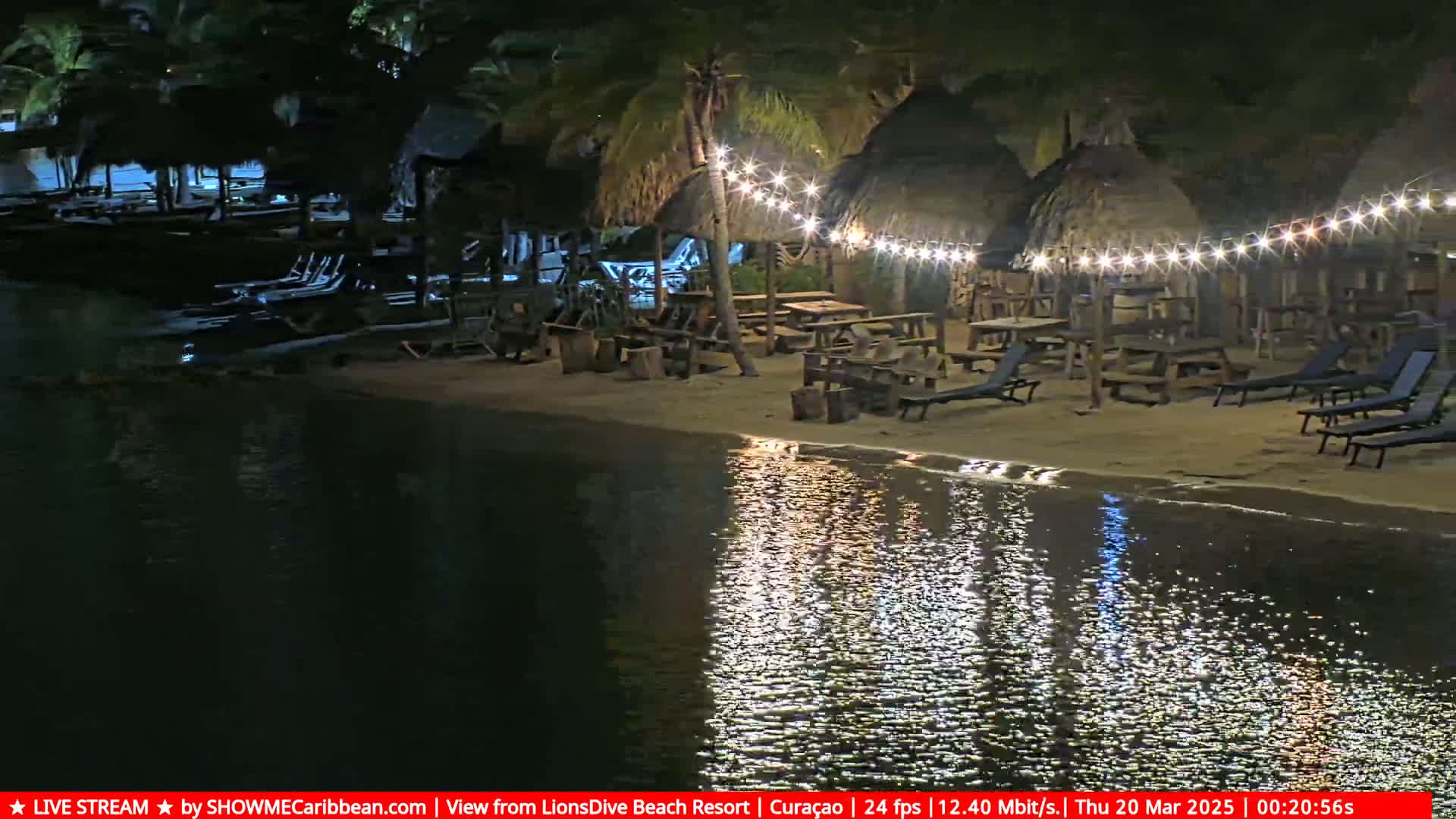 A nighttime beach scene shows several thatched-roof structures with string lights illuminating picnic tables and lounge chairs along the water's edge, with calm water reflecting the lights.