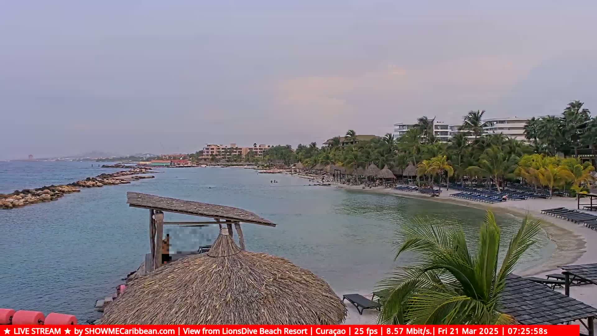 A calm, overcast day reveals a tranquil beach scene with a sandy shore, turquoise water, palm trees, beach chairs, and resort buildings in the background.