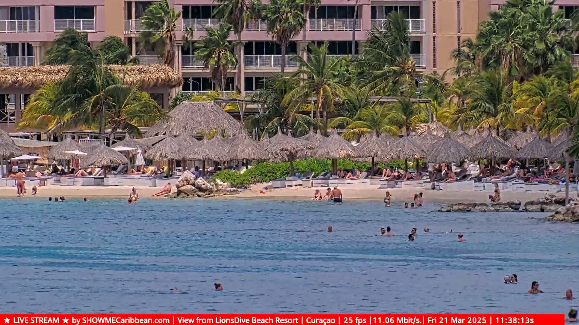 A sandy beach with numerous thatched umbrellas and sunbathers is situated in front of a multi-story building; people are swimming in calm, clear turquoise water.