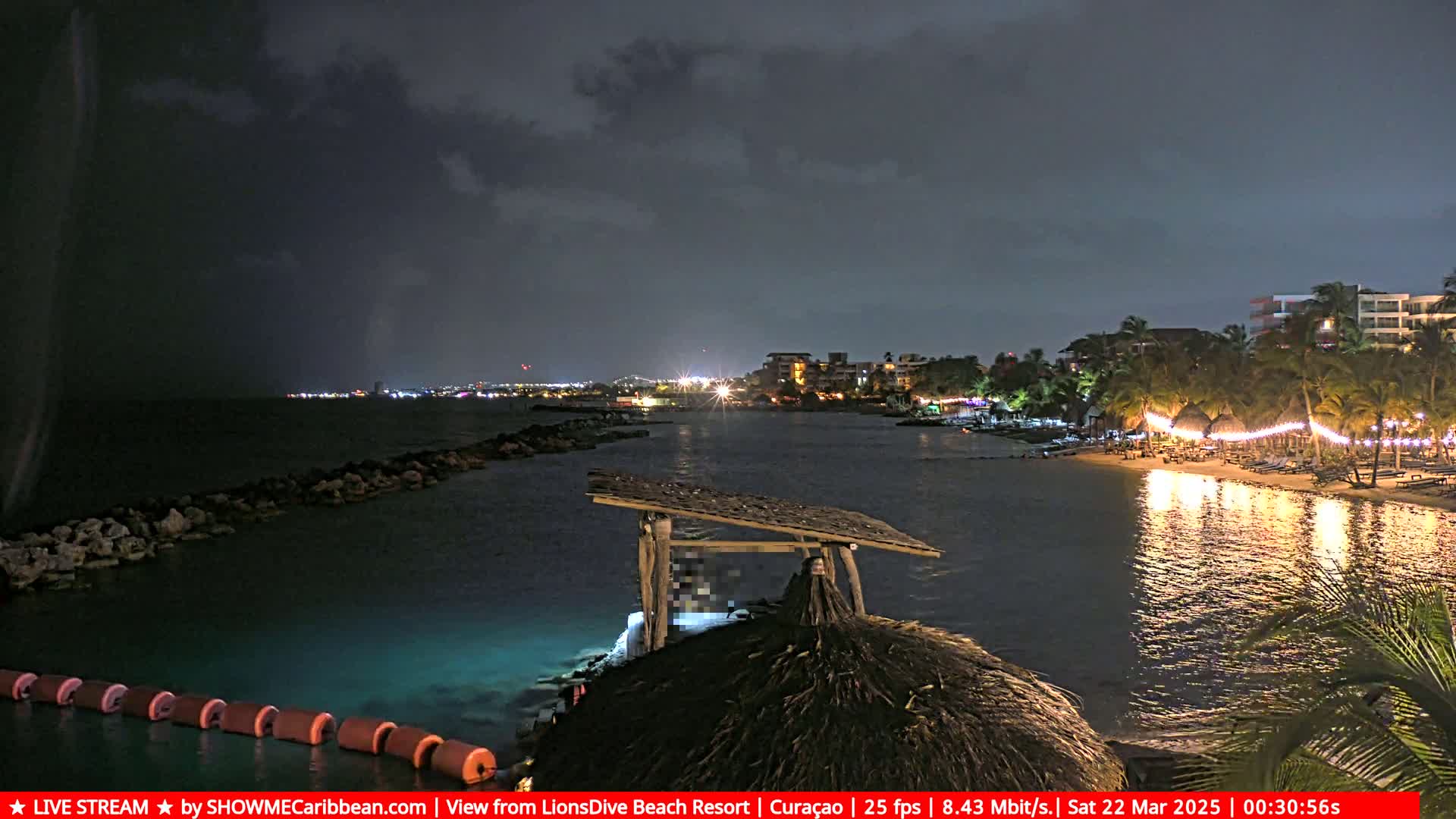 A nighttime beach scene under a dark, cloudy sky shows a calm body of water with lights illuminating a shoreline, buildings in the distance, and a partially obscured thatched structure in the foreground.
