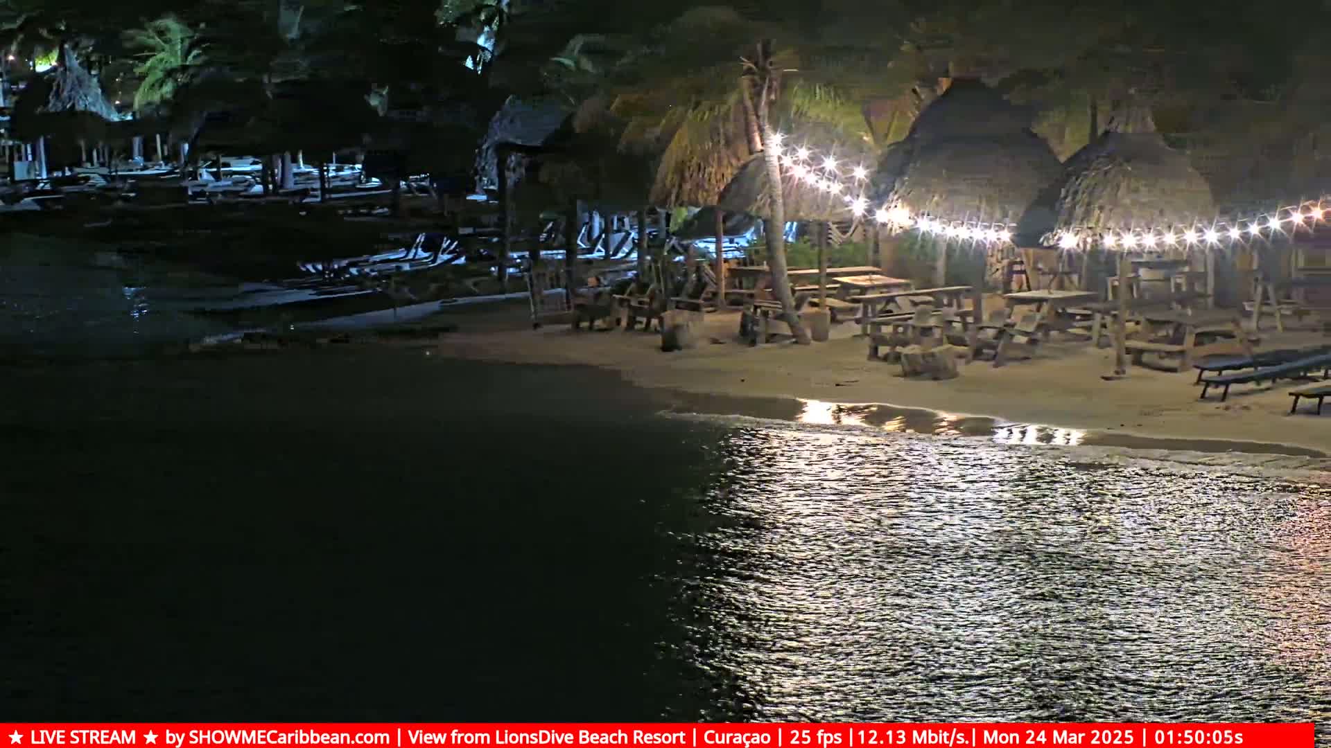 A nighttime beach scene shows several thatched-roof huts with string lights illuminating picnic tables and lounge chairs on a sandy beach next to calm water.