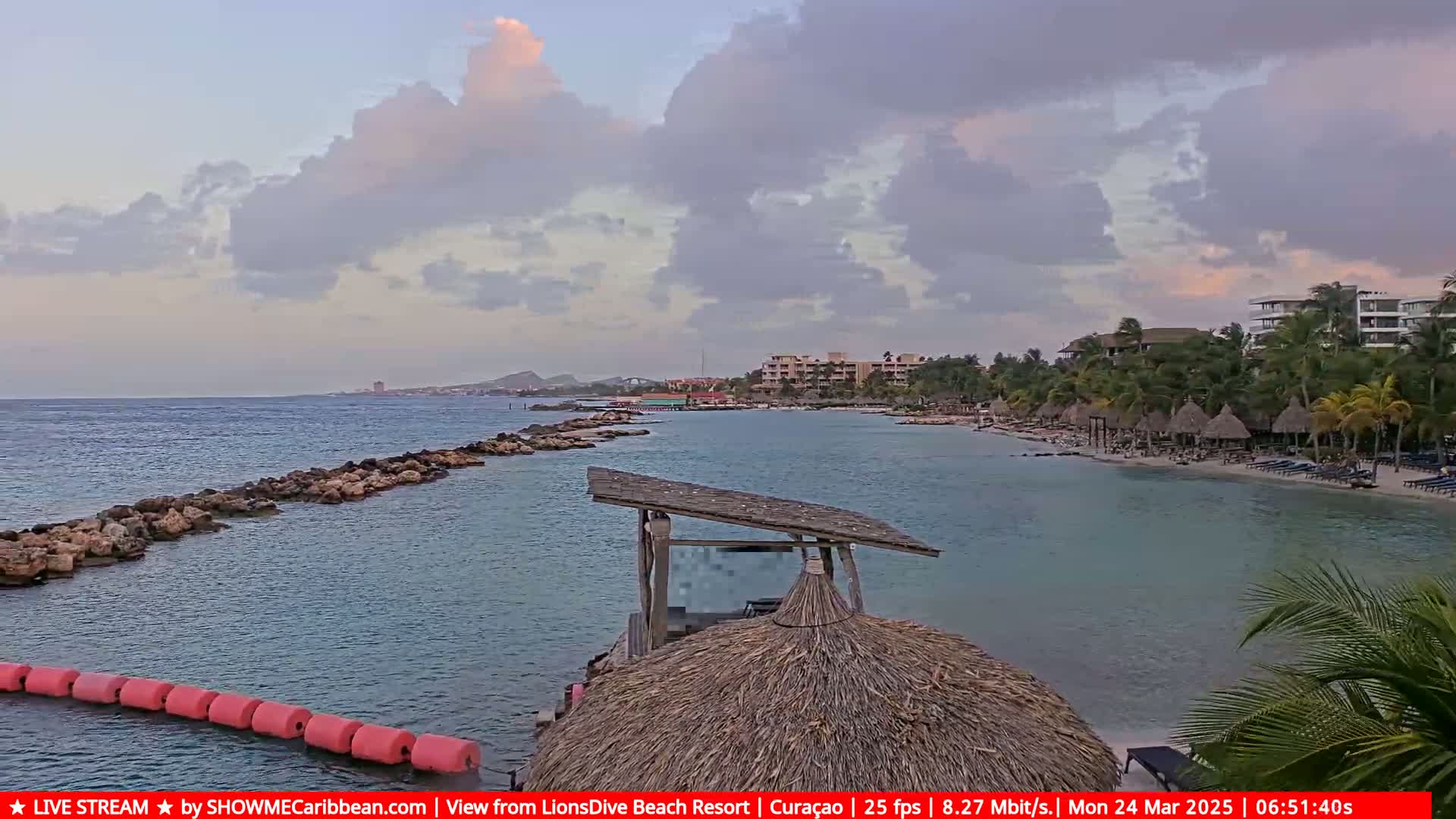 A partly cloudy day shows a tranquil beach scene with calm, clear water, a rocky jetty, beach umbrellas, and resort buildings.