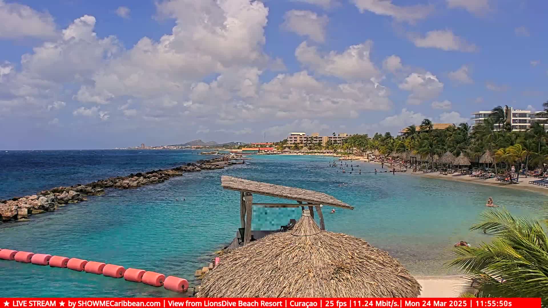 A partly cloudy day at a tropical beach with calm, turquoise water, a sandy shore lined with palm trees and beach umbrellas, and resort buildings in the background.