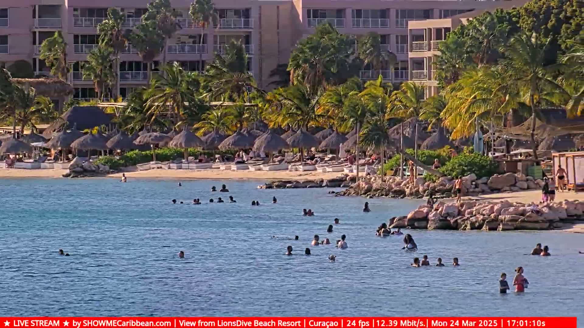 A sunny beach scene shows many people swimming and relaxing in calm, clear water near a sandy shore lined with thatched umbrellas, palm trees, and a resort building.