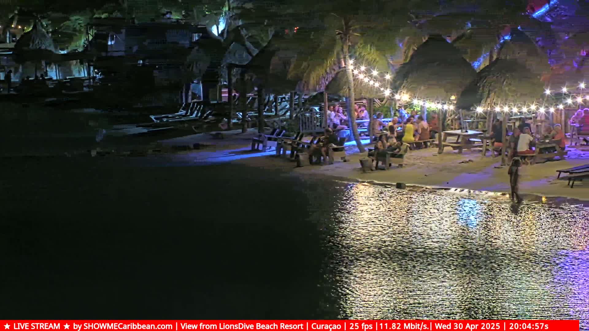 A nighttime beach scene shows people gathered at picnic tables under thatched-roof structures lit with string lights, beside calm, dark water reflecting the lights.