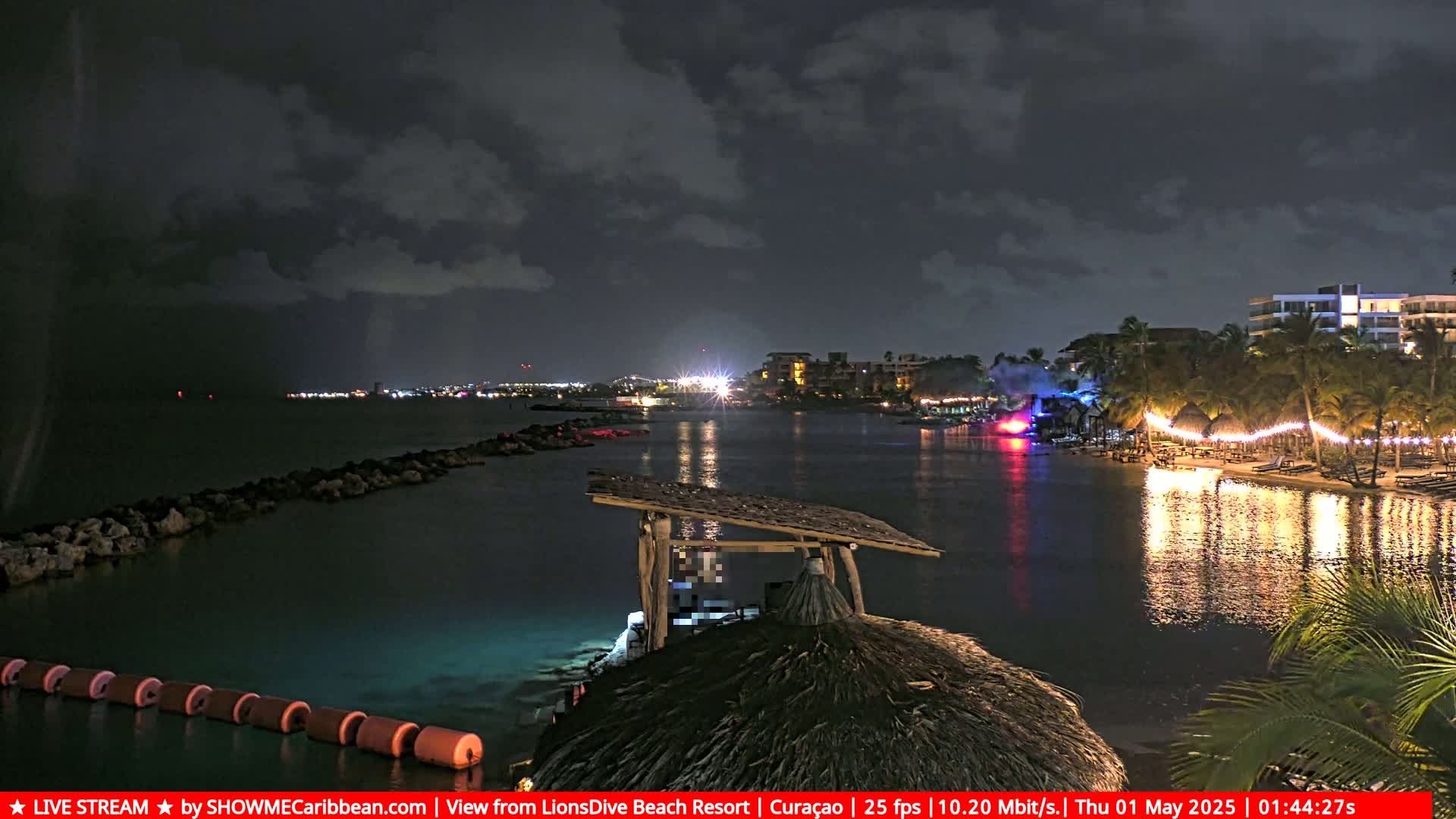 A nighttime view of a calm bay with a resort's lights reflecting on the water under a partly cloudy sky.