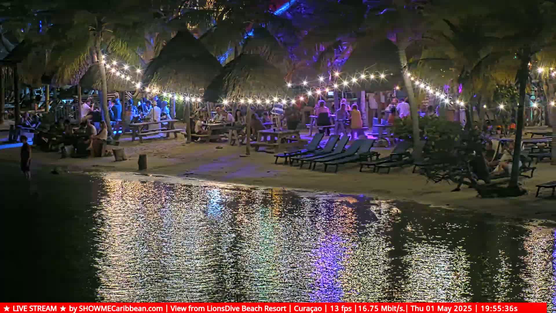 A nighttime beach scene shows people gathered at picnic tables and lounge chairs under thatched-roof structures illuminated with string lights, reflected in the calm water.
