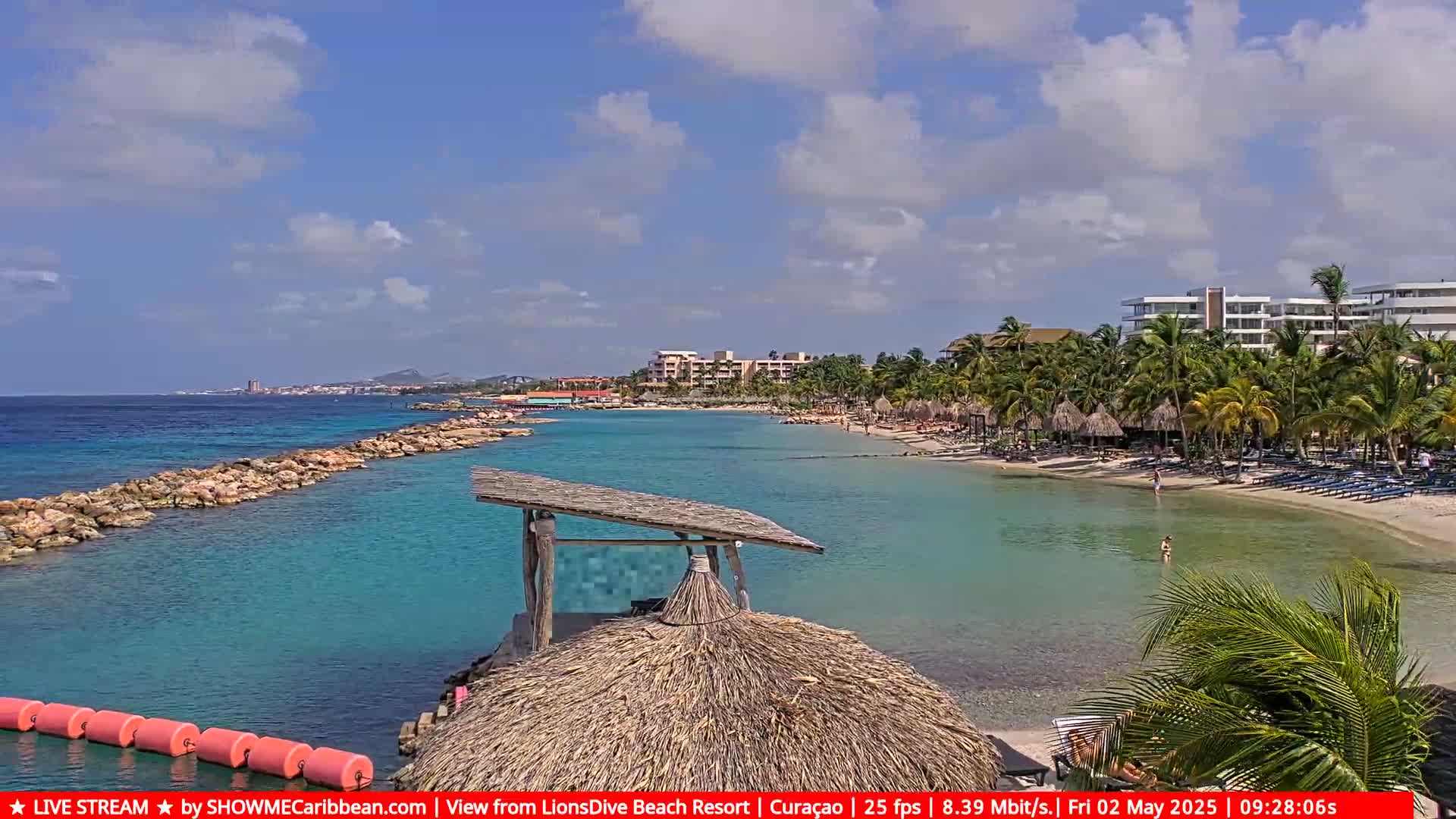 A calm, sunny day at a tropical beach with turquoise water, a rocky breakwater, palm trees, beach chairs, and resort buildings in the background.