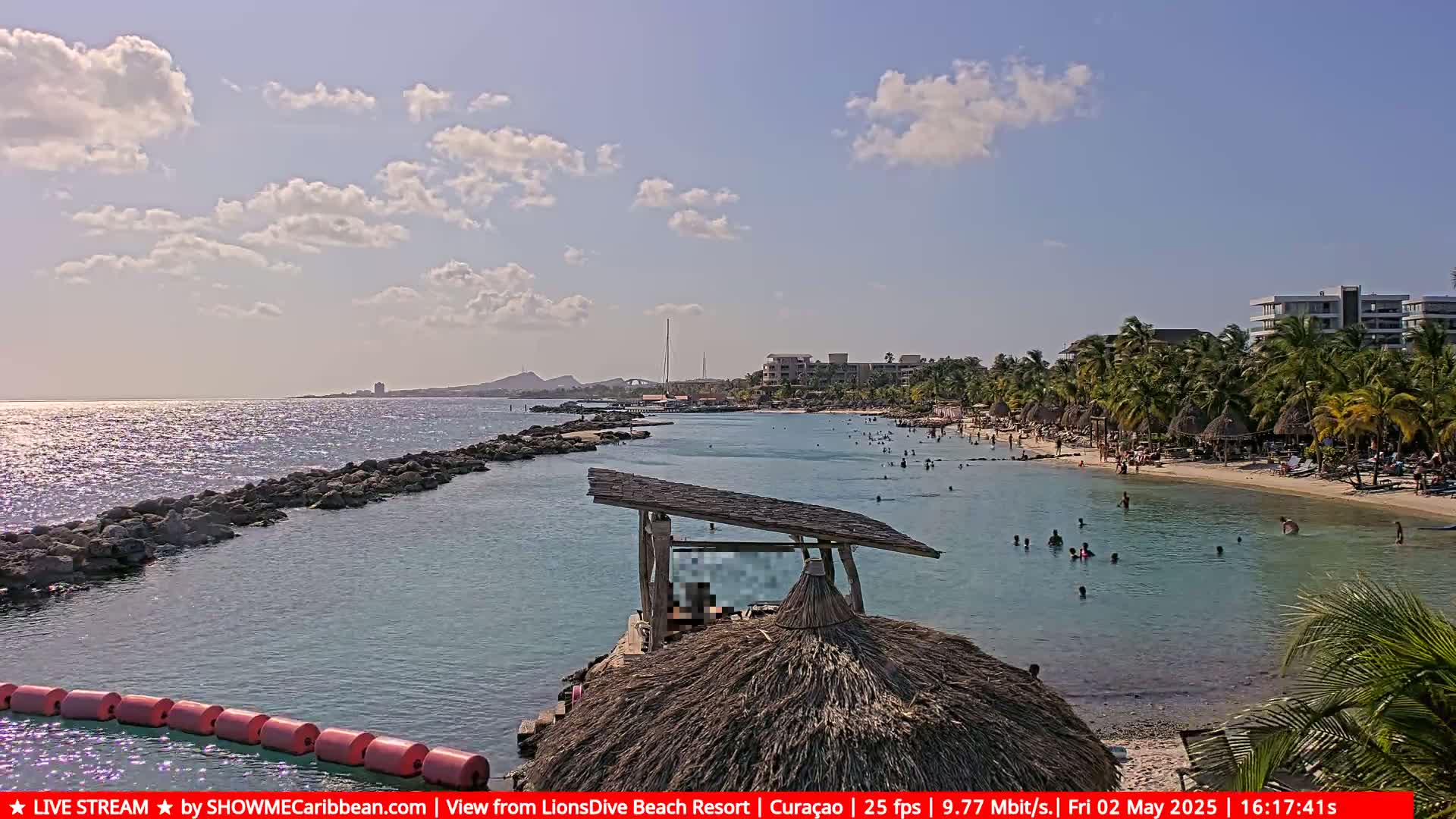 A partly sunny day at a tropical beach shows calm turquoise water, a sandy shore with beachgoers, palm trees, and resort buildings in the background.
