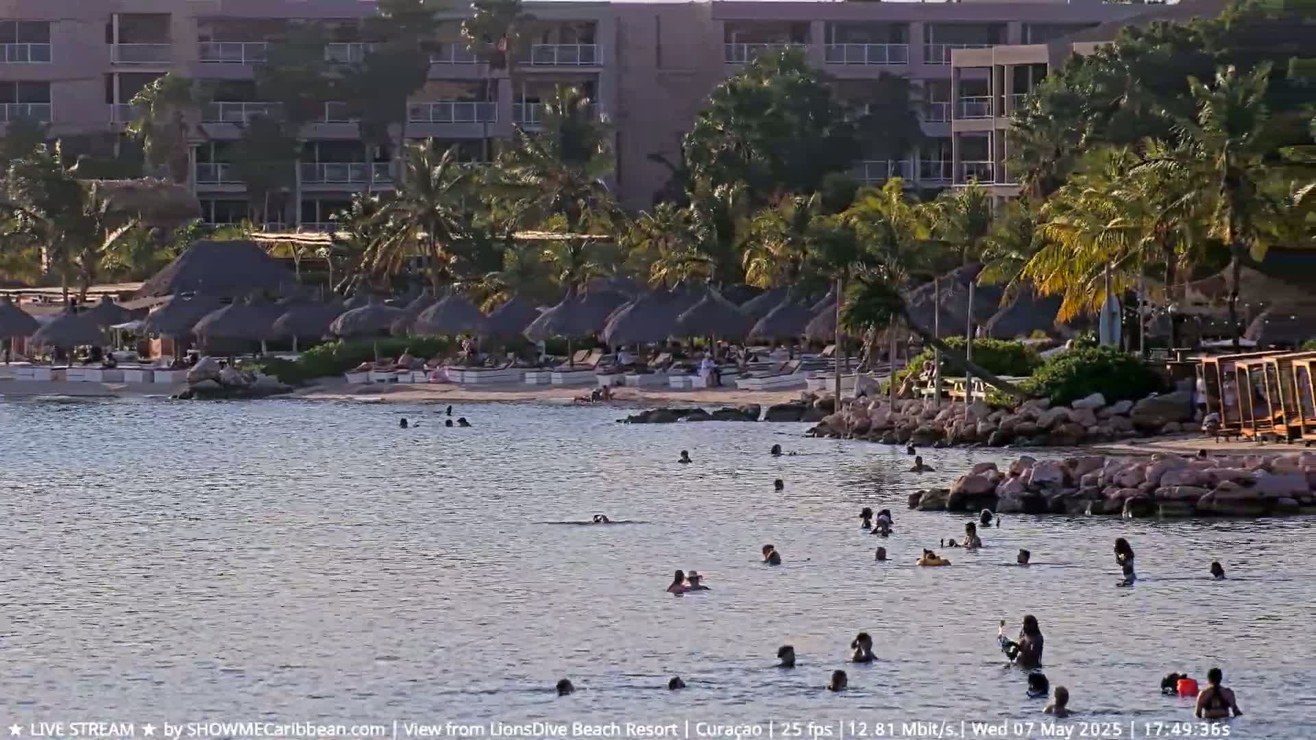 A calm body of water with many people swimming near a sandy beach lined with thatched-roof umbrellas and a resort building in the background.