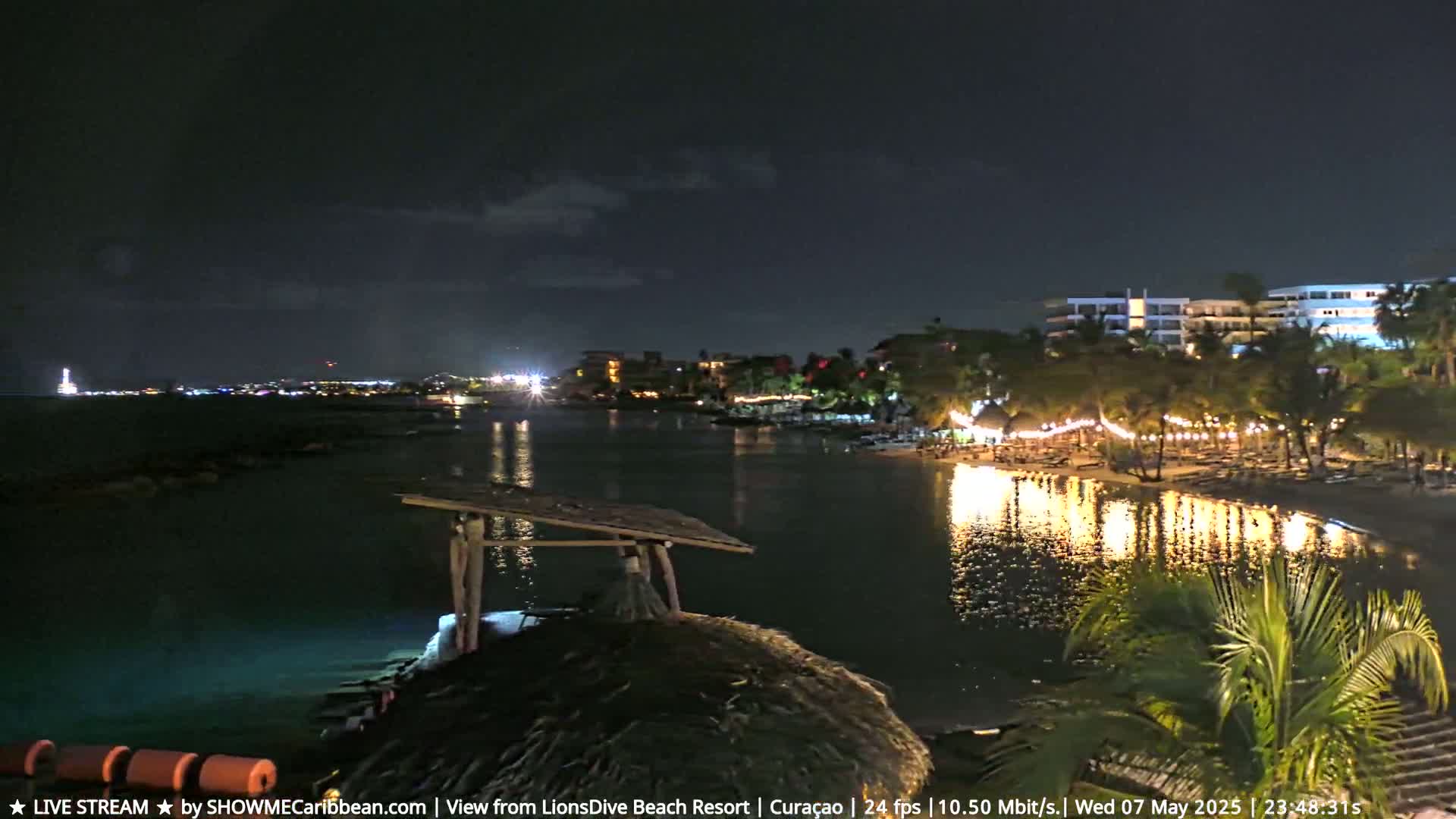 A nighttime view of a calm, illuminated body of water reflecting lights from a beach resort and buildings across the water under a mostly clear sky.
