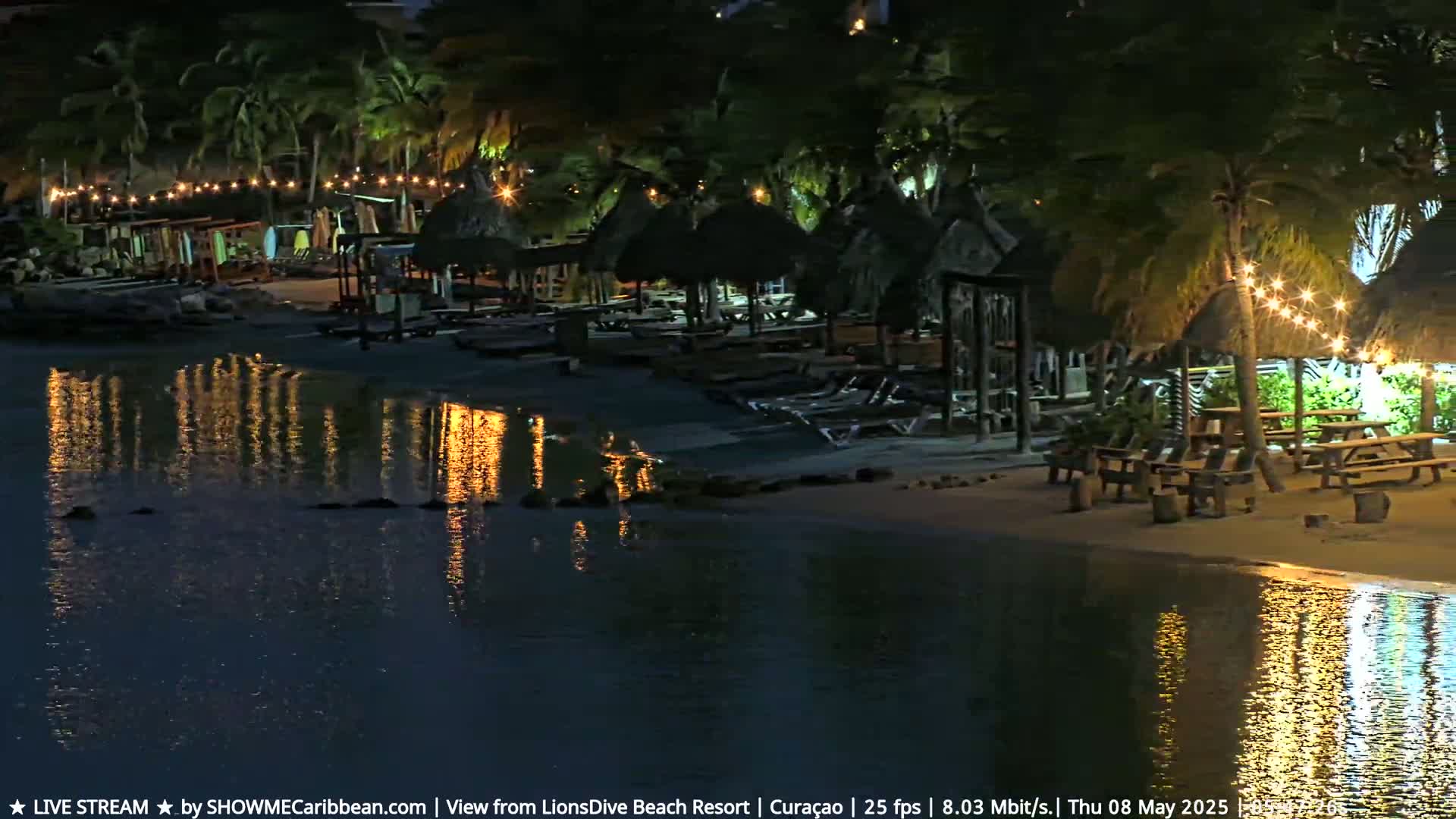 A nighttime beach scene shows calm water reflecting the lights of several thatched-roof beach huts and picnic tables lined along the shore, under a dark sky.